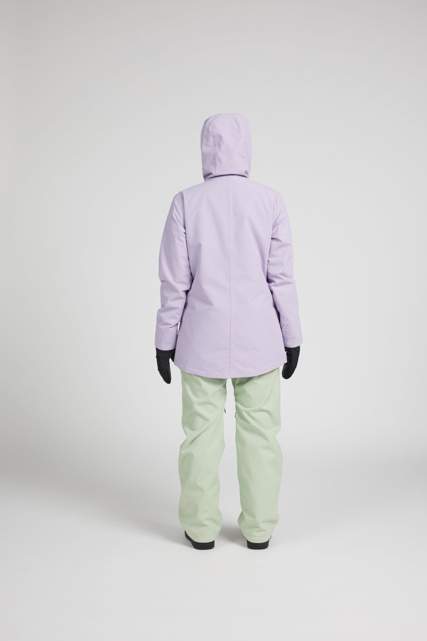 A person shown from the back wearing a light lavender hooded jacket, mint green snow pants, and black gloves, standing against a plain light gray studio background.
