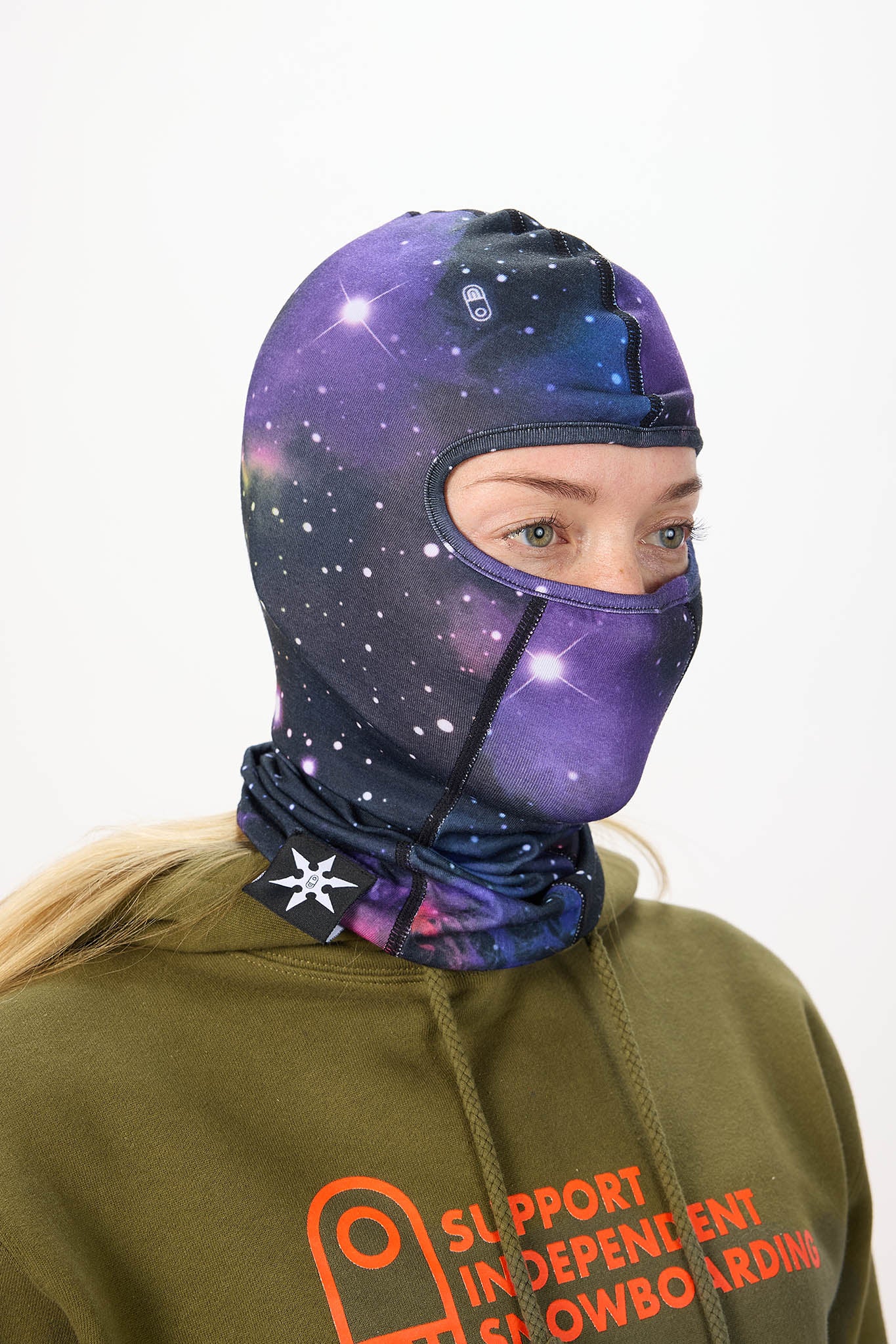 A person wearing a full-head space-themed balaclava mask with a purple and blue galaxy print, paired with an olive green hoodie against a plain white background.