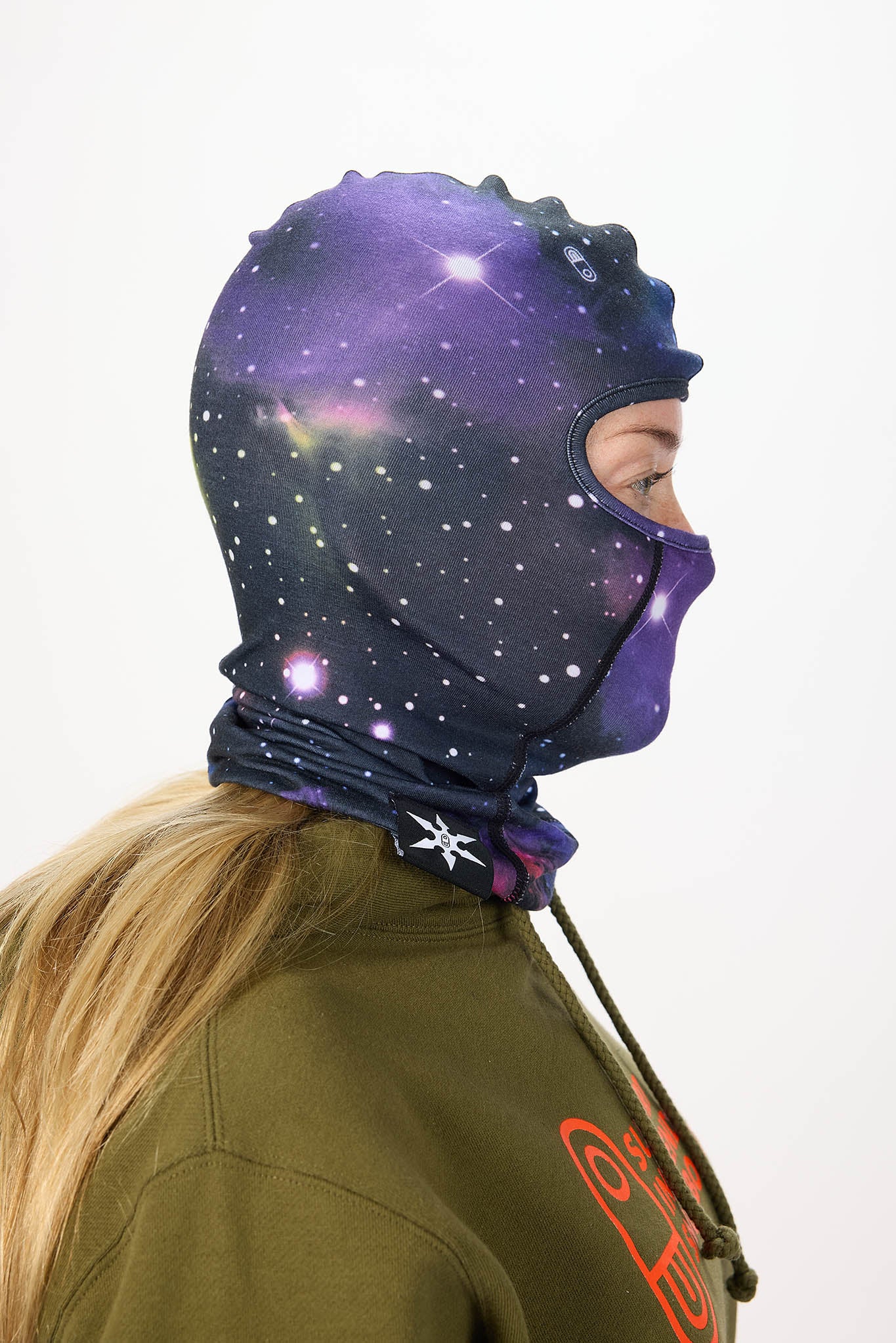 A person shown in side profile wearing a full-head space-themed balaclava hood with a purple and blue galaxy print, paired with an olive green hoodie against a plain white background.