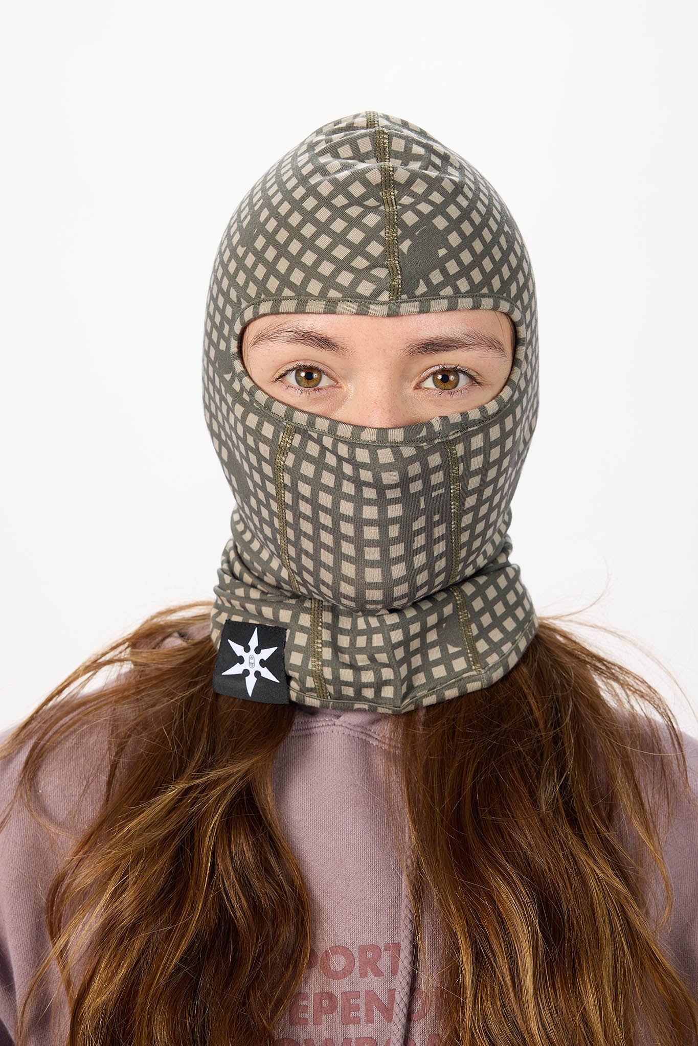 A person wearing a gray and beige patterned balaclava face mask with a black logo patch on the chin, shown against a plain white background.
