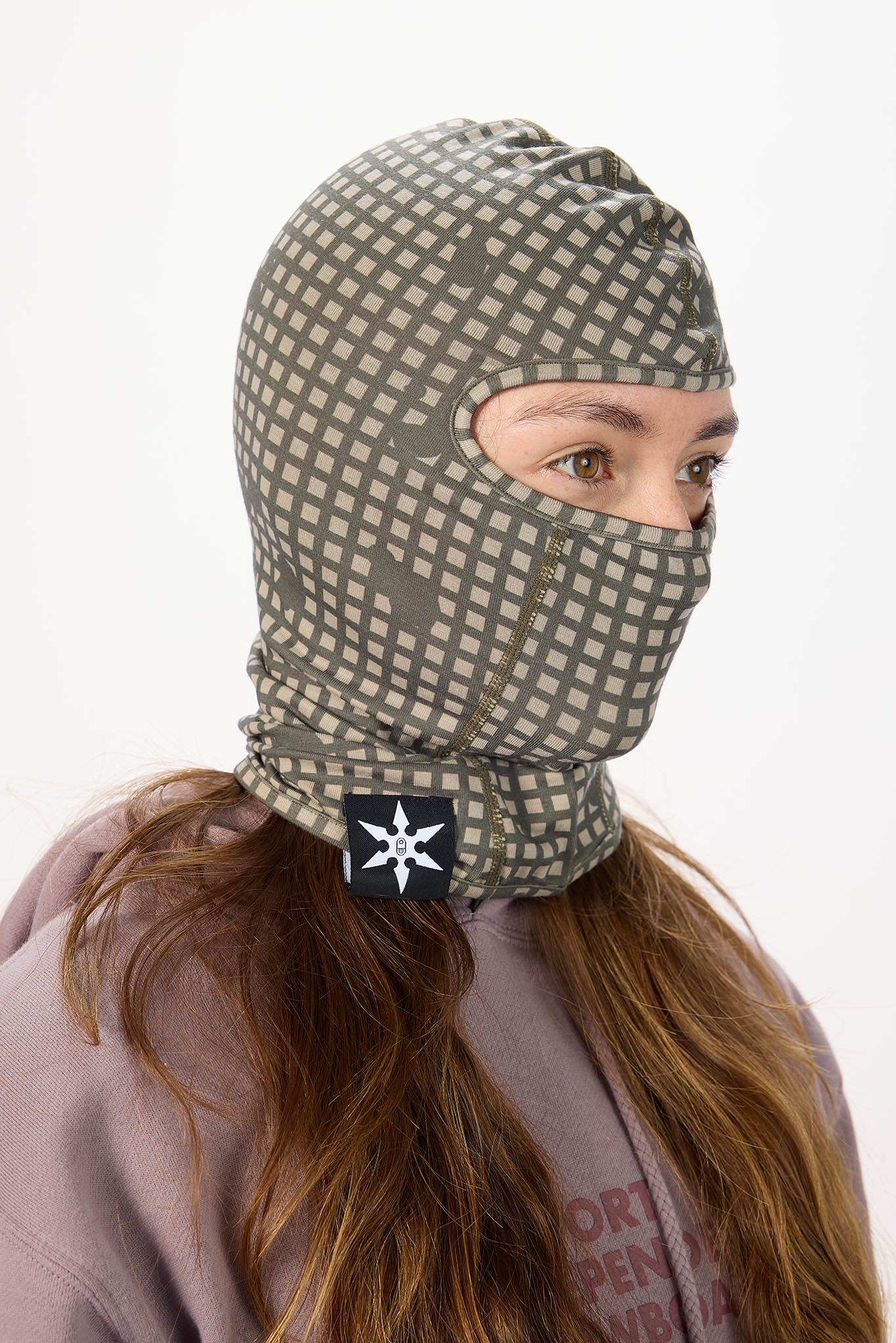 A person wearing a gray and beige patterned balaclava face covering with a black patch featuring a white compass star logo, set against a plain white background.