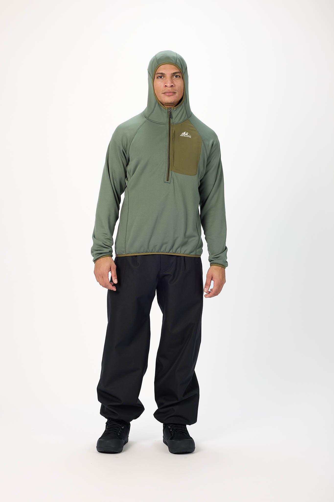 A man modeling a sage green hooded quarter-zip pullover with an olive green chest pocket and black pants, standing against a plain white studio background.