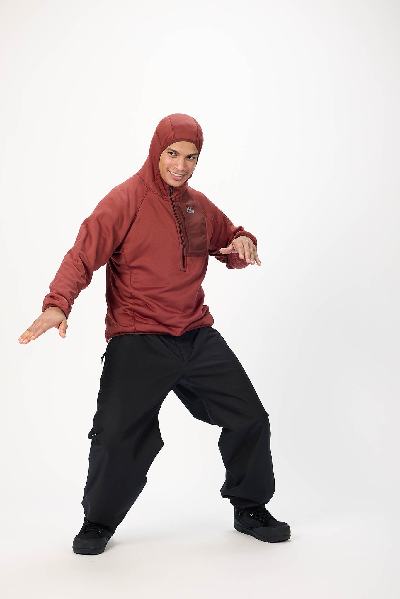 A man posing in a studio wearing a rust-red hooded zip-up jacket and black cargo pants against a white background.