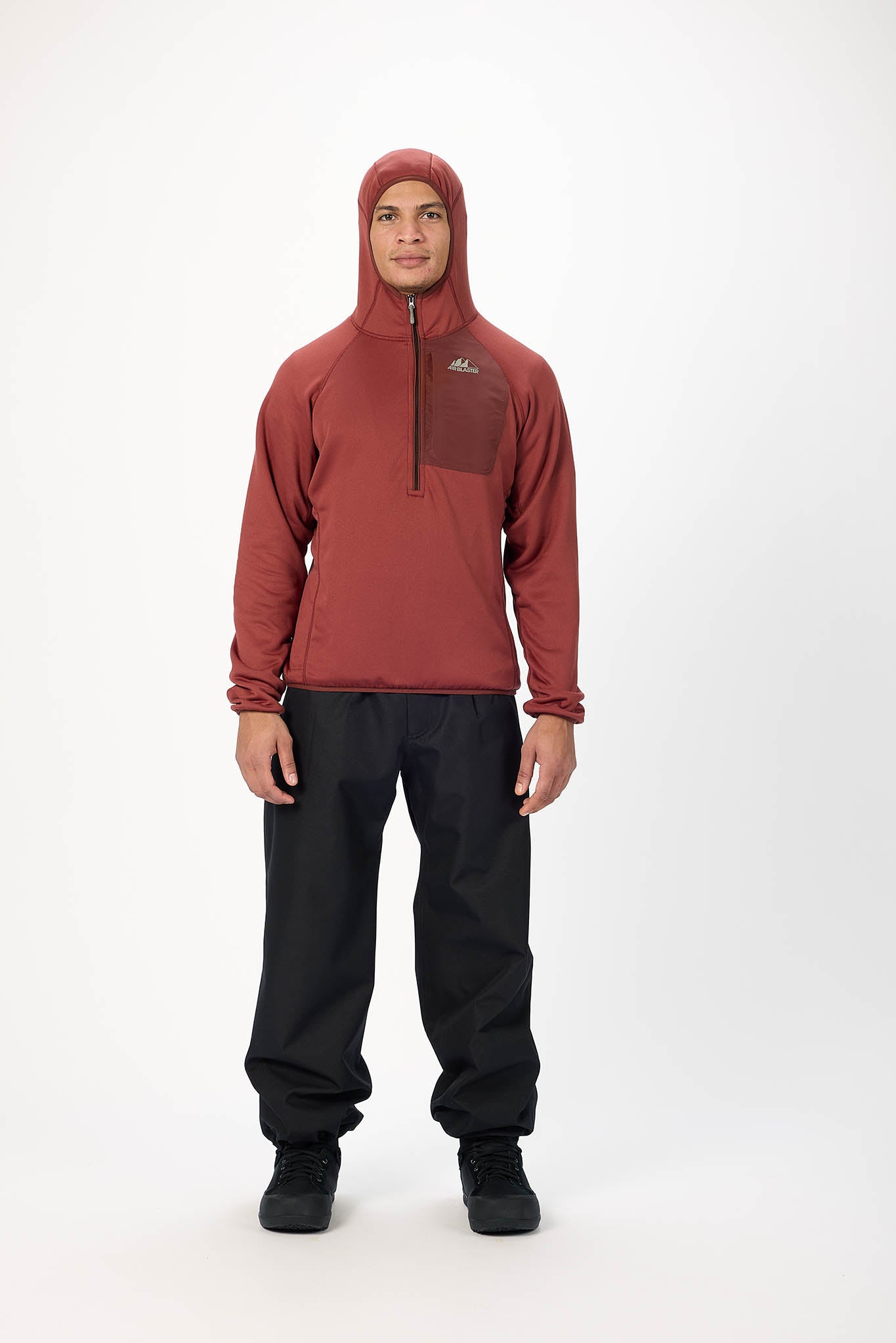 A man wearing a rust-colored hooded quarter-zip pullover jacket and black pants, standing against a plain white studio background.