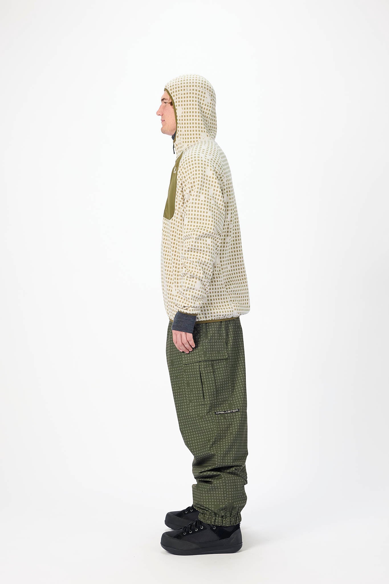 A person shown in side profile wearing a cream and olive patterned hooded jacket, olive green pants, and black shoes, photographed against a plain light gray studio background.