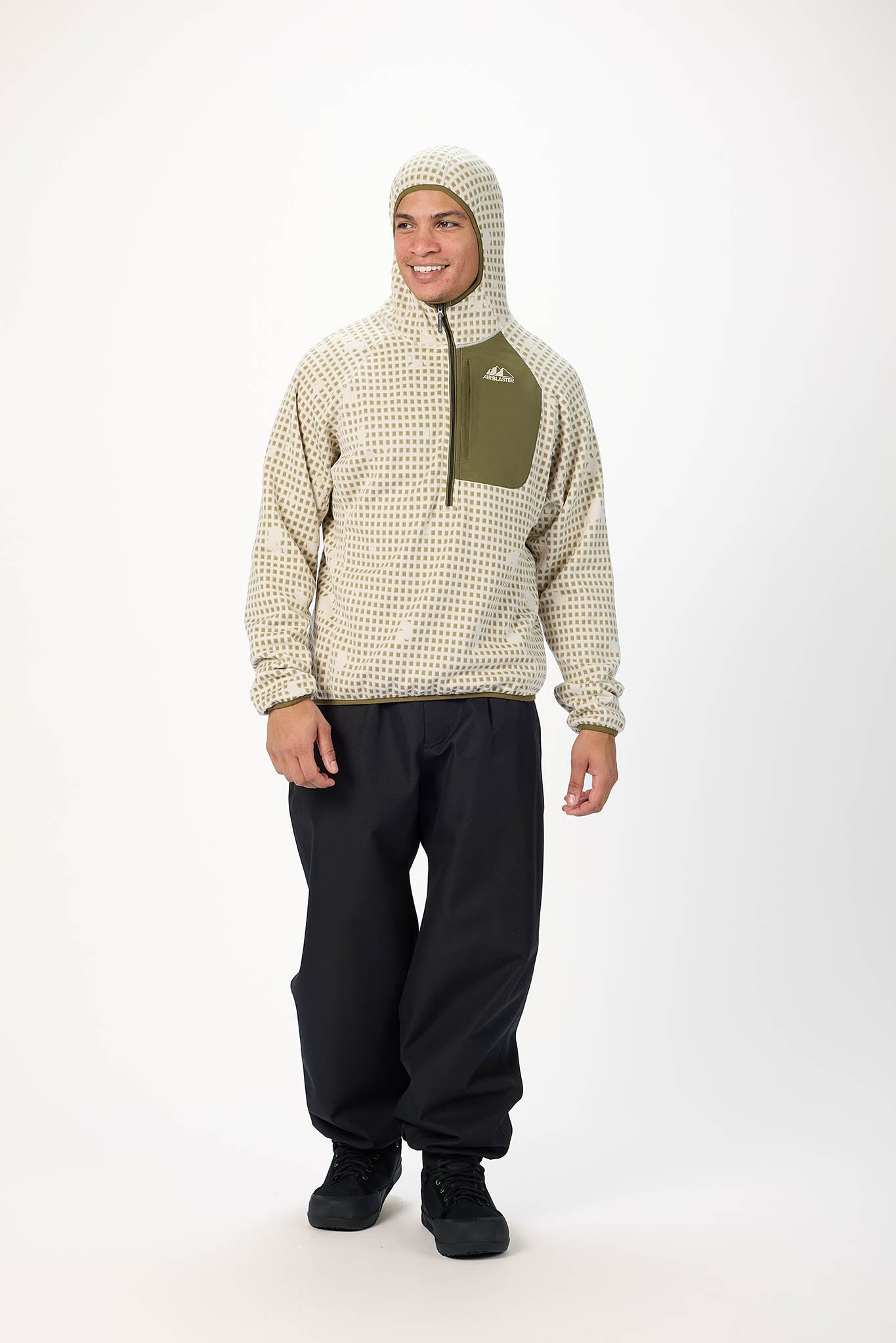 A man modeling a fleece pullover hoodie with a beige and white micro-check pattern, olive green chest pocket, and matching patterned hood, paired with black pants and black boots, against a plain white studio background.
