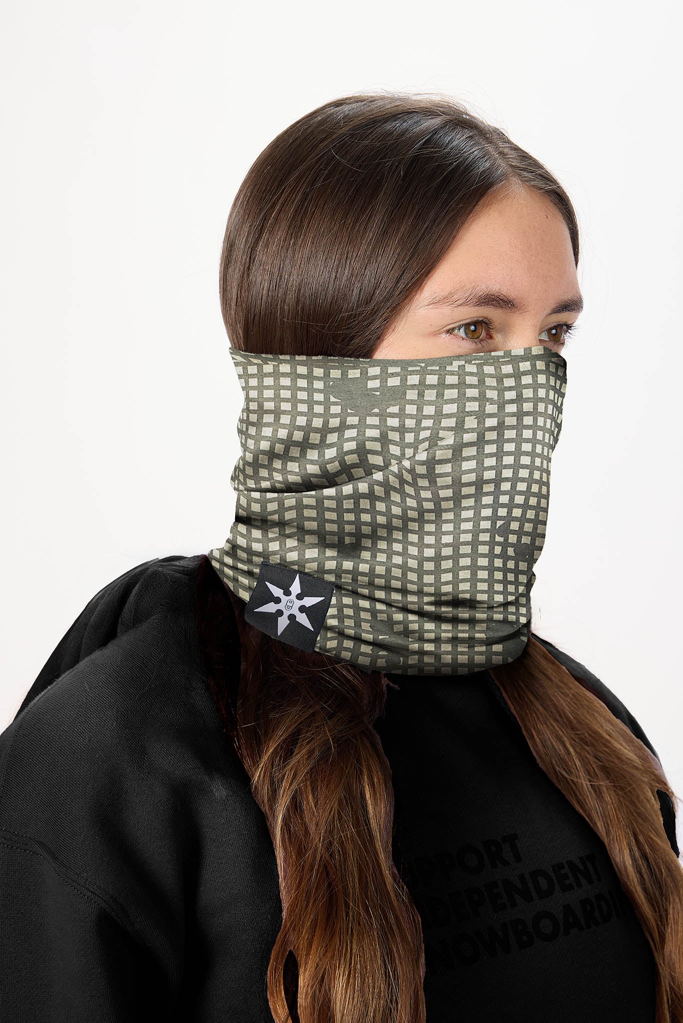 A woman wearing a patterned face gaiter neck warmer in olive green and white, paired with a black hoodie against a plain white background.