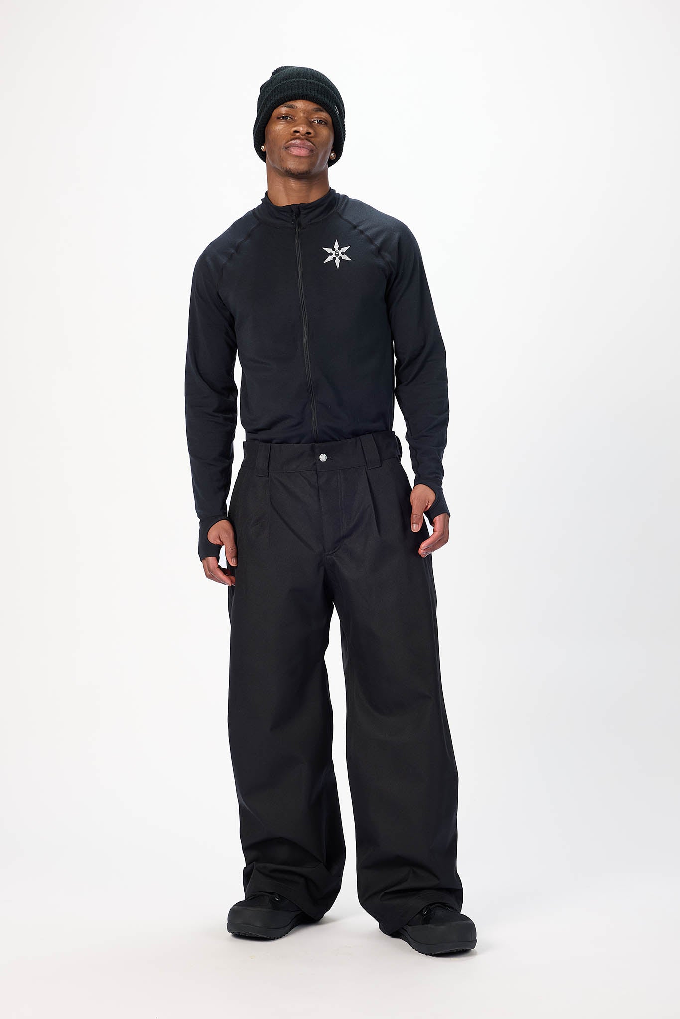 A model wearing a black full-body tracksuit with a white geometric logo on the chest, a black beanie, and black shoes, standing against a plain white studio background.