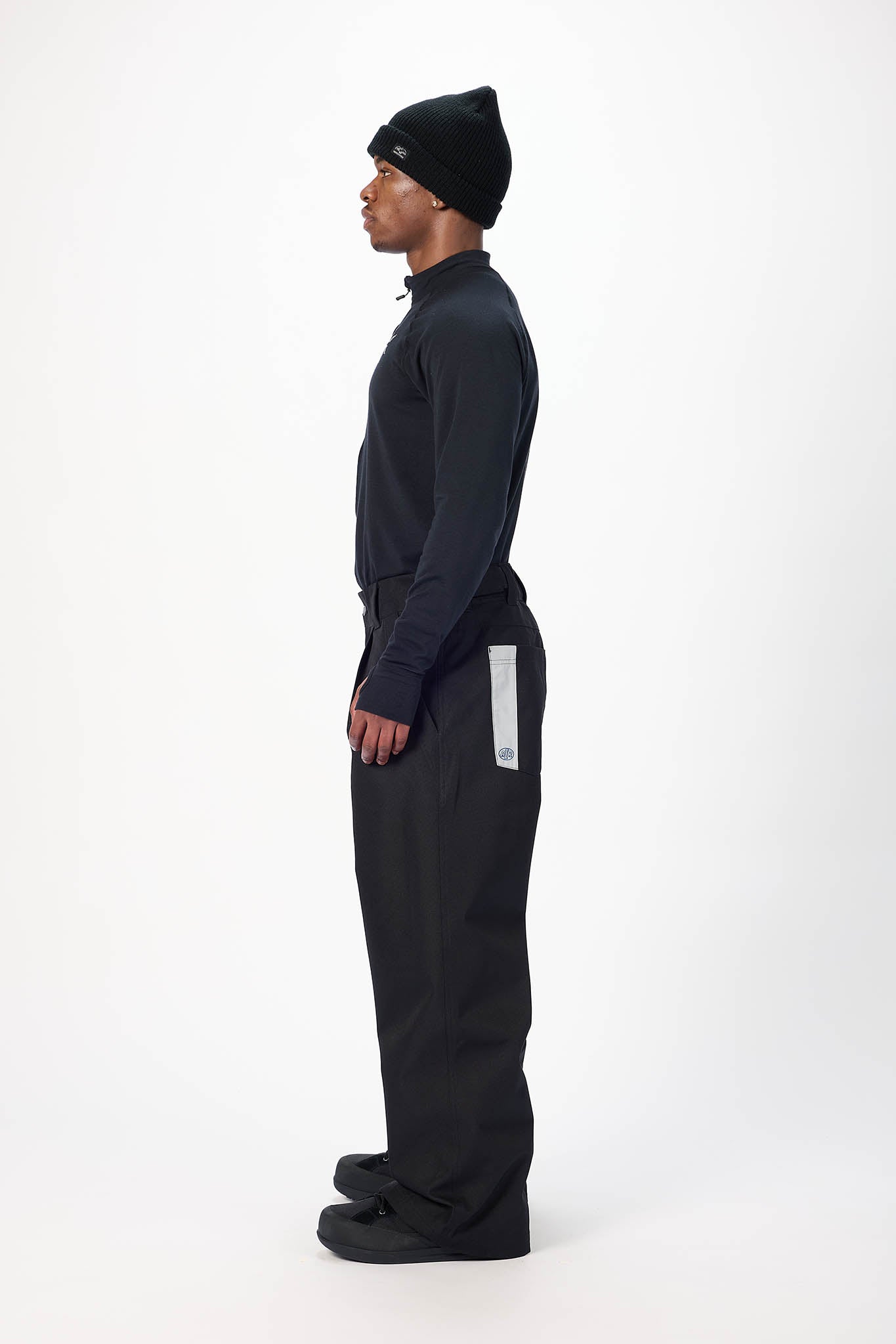 A model shown in profile wearing a black knit beanie, a black long-sleeve mock-neck top, and black wide-leg pants with a reflective white vertical stripe on the side, standing against a plain white studio background.