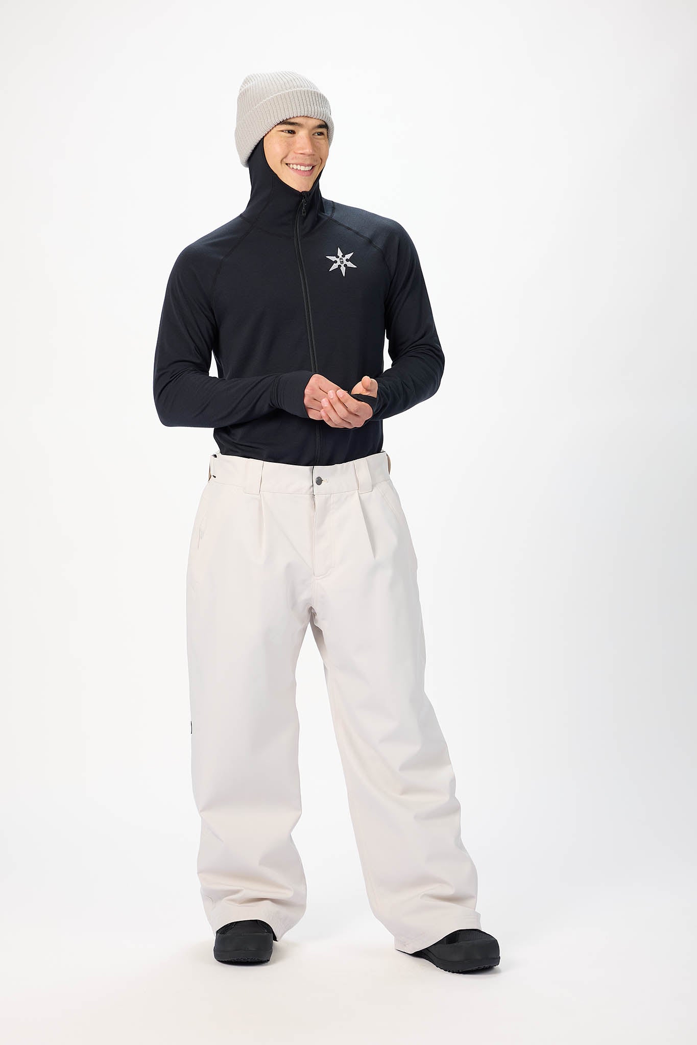 A man modeling a black long-sleeve zip-up jacket with a white star logo on the chest, paired with off-white snow pants, a light gray beanie, and black shoes, against a plain white studio background.