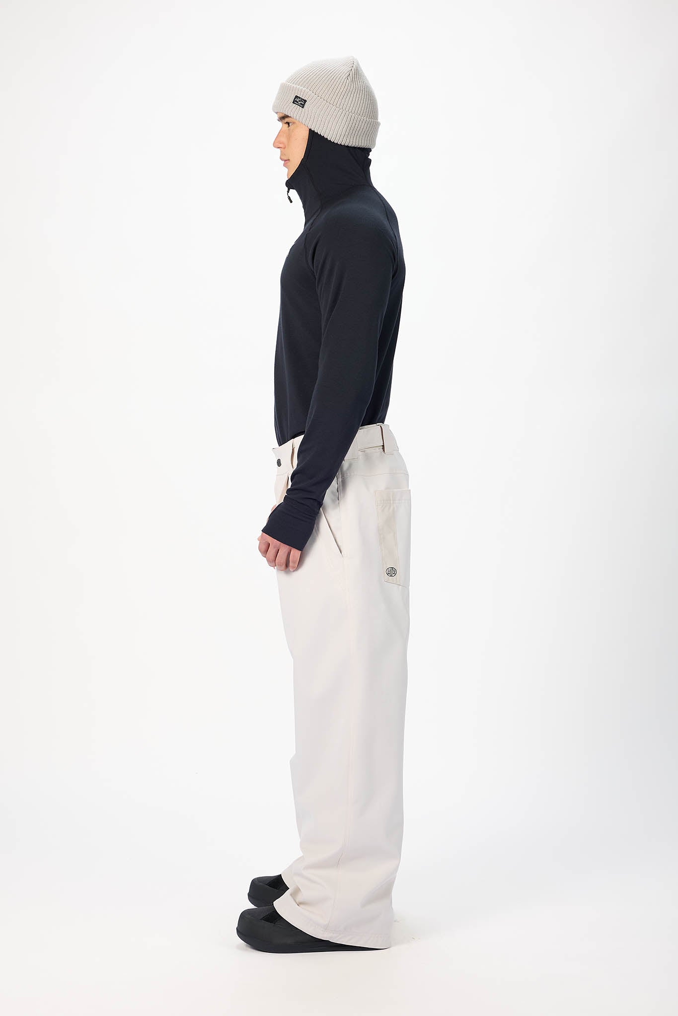 A model shown in profile wearing a black long-sleeve mock-neck top, off-white wide-leg pants, and a light gray beanie against a plain white studio background.