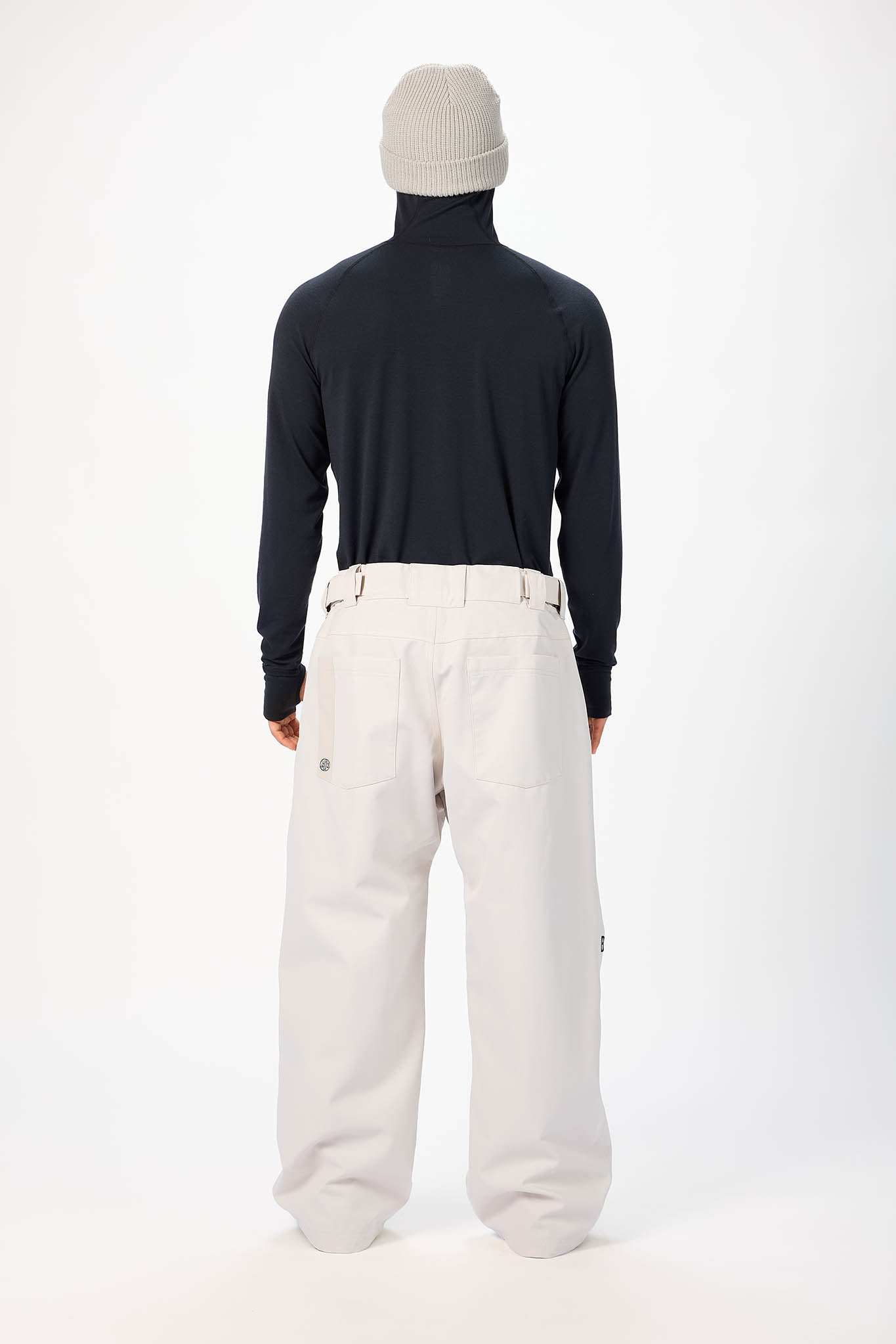 A model shown from the back wearing a black long-sleeve turtleneck, off-white wide-leg snow pants, and a light gray beanie against a plain white studio background.