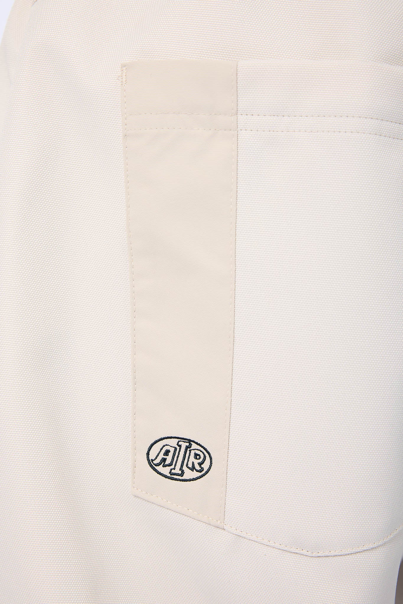 Close-up of a light beige pocket on a white garment, featuring a vertical beige stripe and a black and white circular 'AIR' logo patch near the bottom.