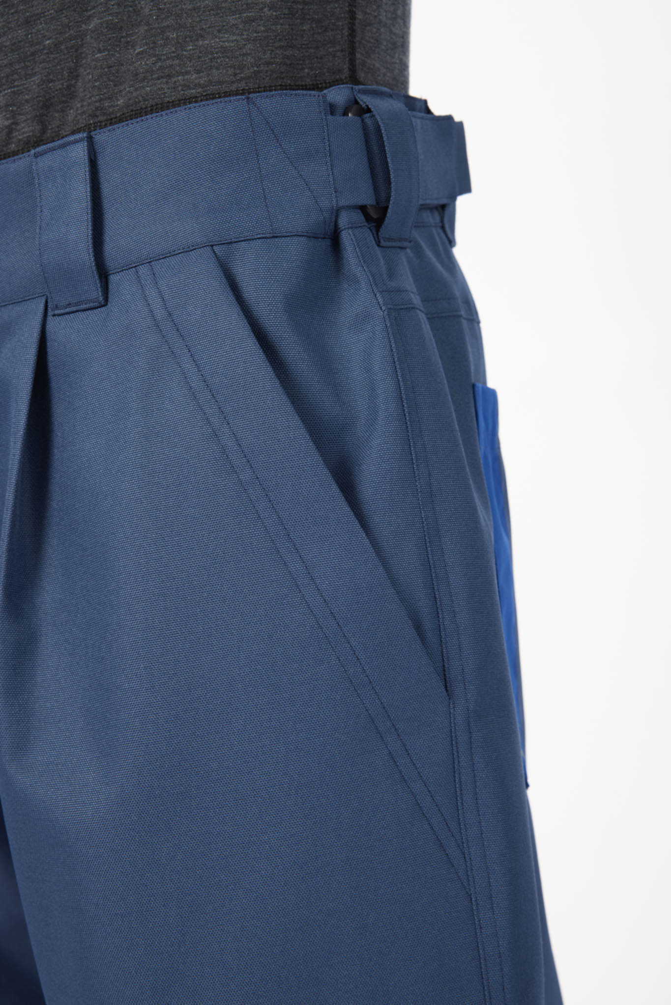 Close-up of a pair of blue pleated trousers with a belt loop and side pocket, shown on a model wearing a dark gray top against a white background.