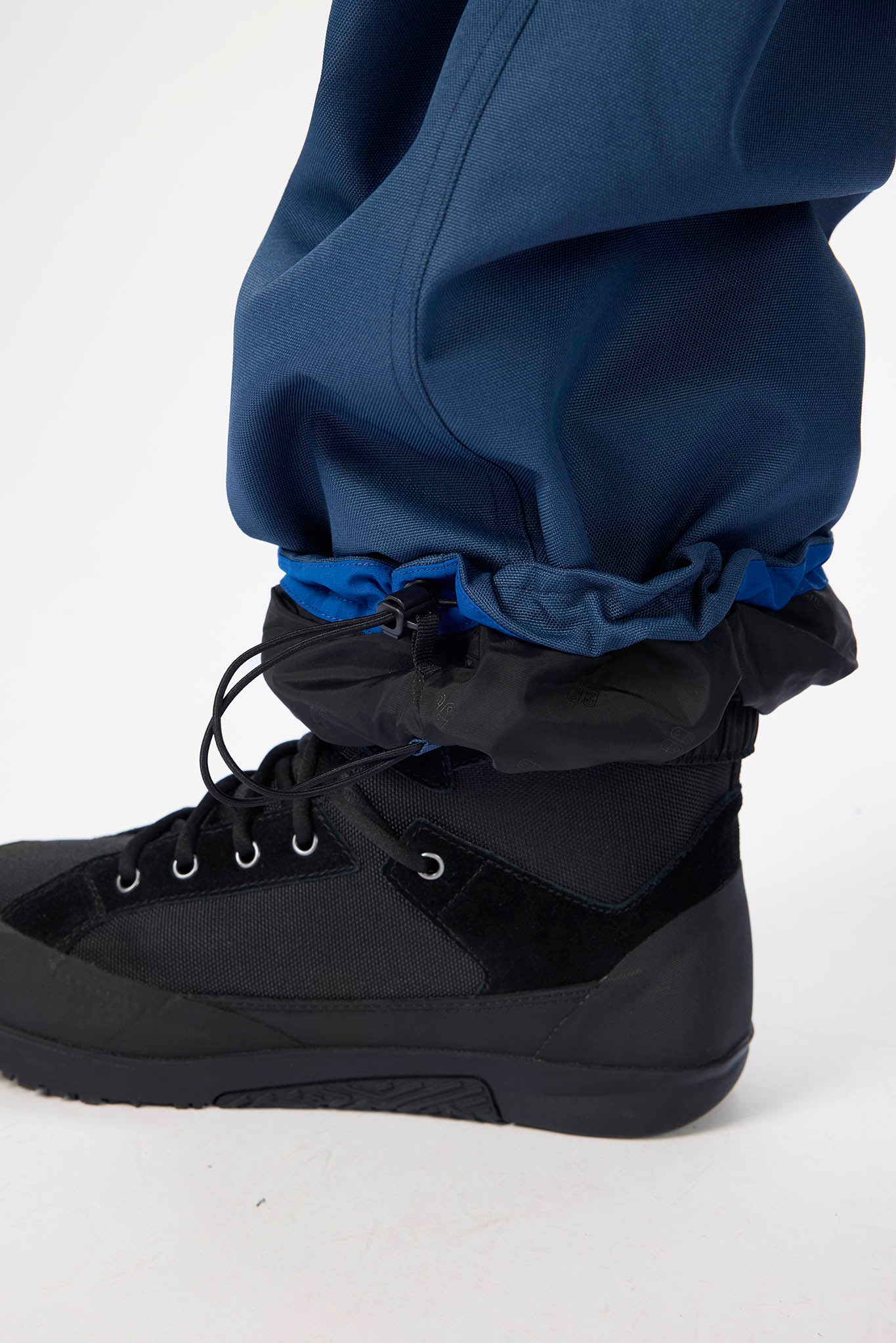 A close-up of a person's lower leg wearing navy blue pants with a black and navy gaiter cuff, paired with a black lace-up boot, on a white studio background.