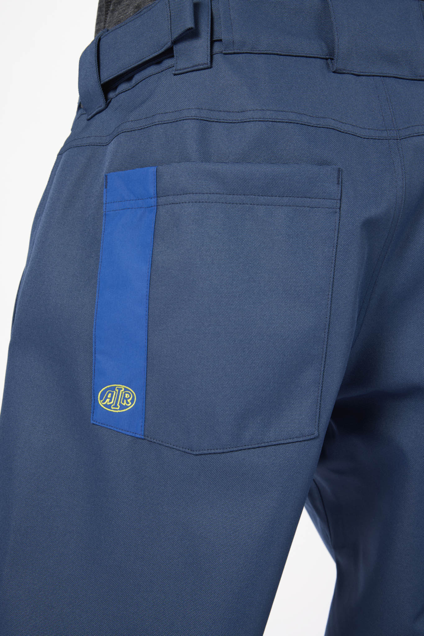 Close-up of the back waistband and right pocket of blue work pants, featuring a vertical blue stripe on the pocket with a small circular logo patch.