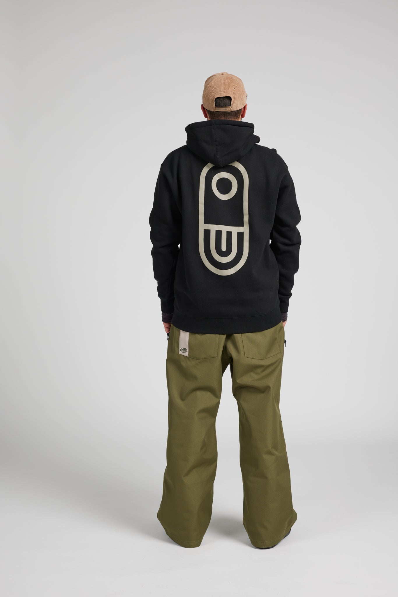 A model shown from the back wearing a black hoodie with a large beige graphic on the back, olive green pants, and a tan baseball cap, set against a plain light gray studio background.
