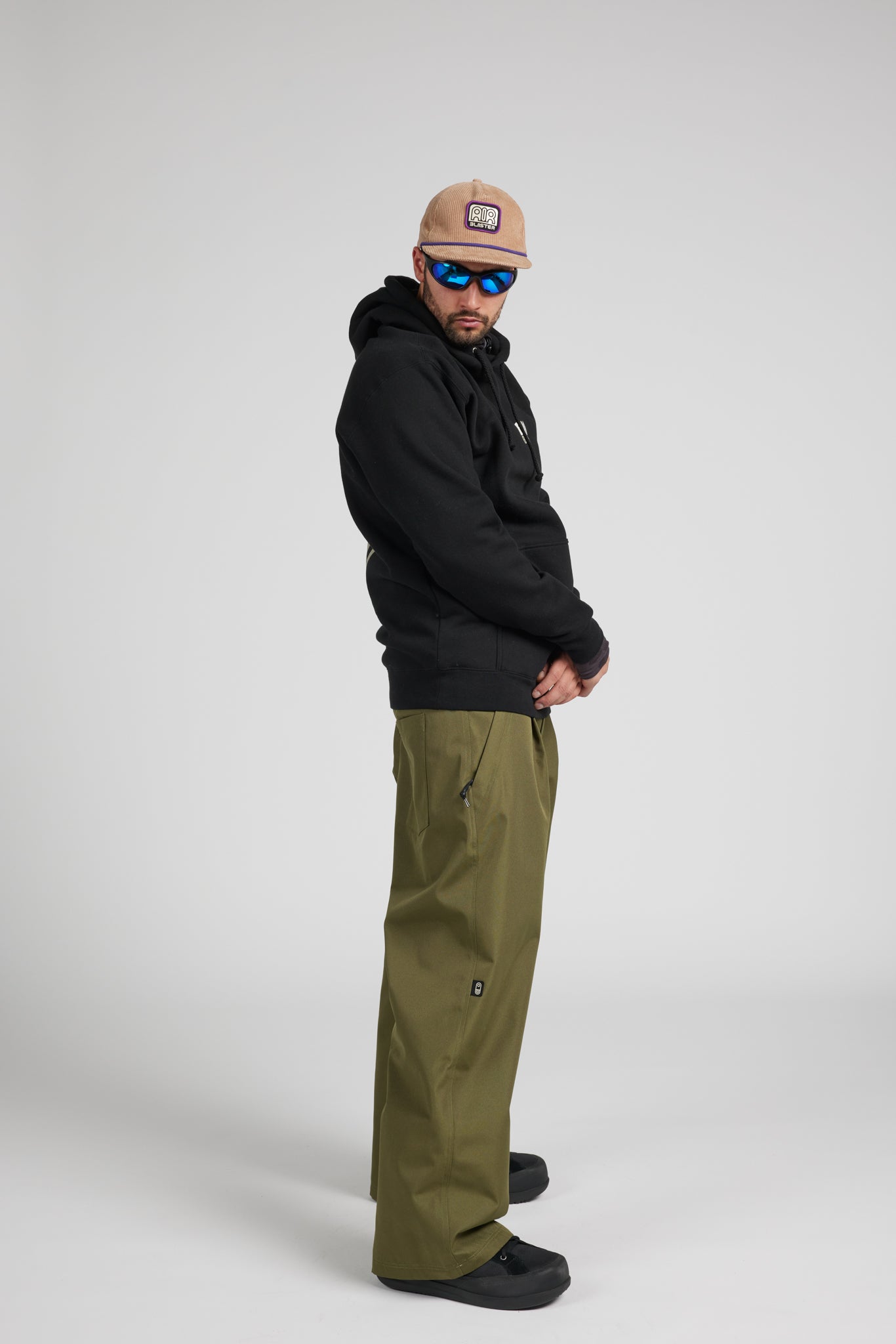 A man wearing a black hoodie, olive green pants, a beige cap, and blue-tinted sunglasses, standing in profile against a light gray studio background.