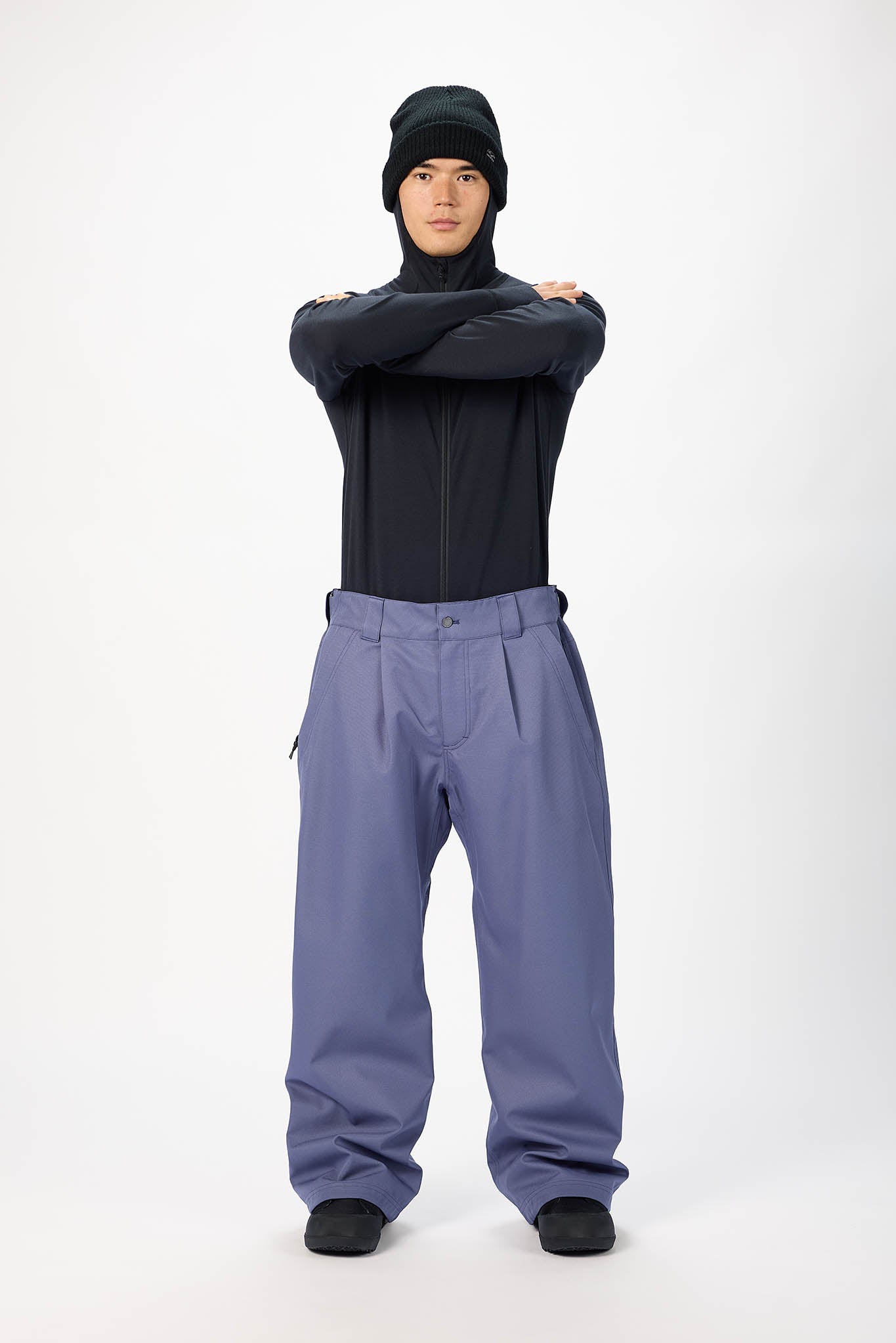A model wearing a black zip-up jacket, a black beanie, and slate blue snow pants, standing with arms crossed against a white studio background.