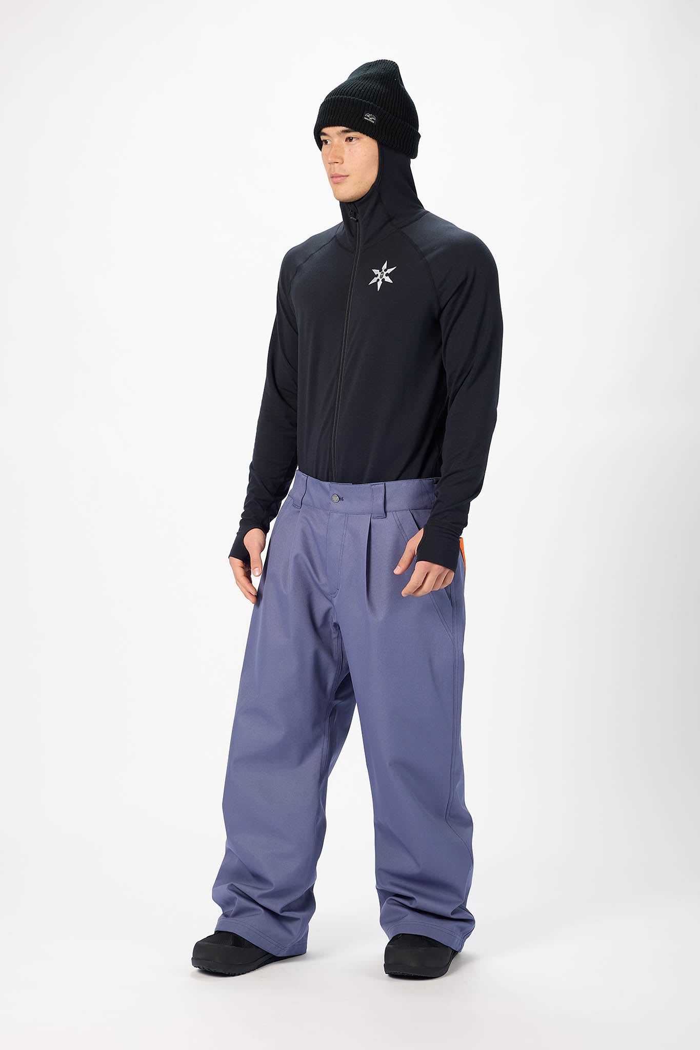 A model wearing a black long-sleeve half-zip jacket with a white logo on the chest, paired with slate blue snow pants, a black beanie, and black gloves, standing against a white studio background.