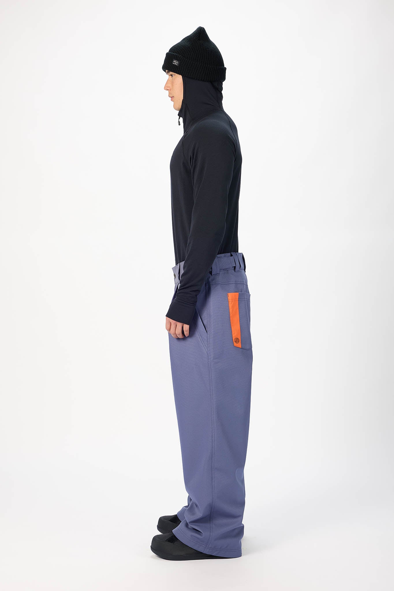 A model shown in profile wearing a black knit beanie, a black long-sleeve top, and wide-leg slate blue pants with a bright orange vertical accent on the side pocket, set against a plain white studio background.
