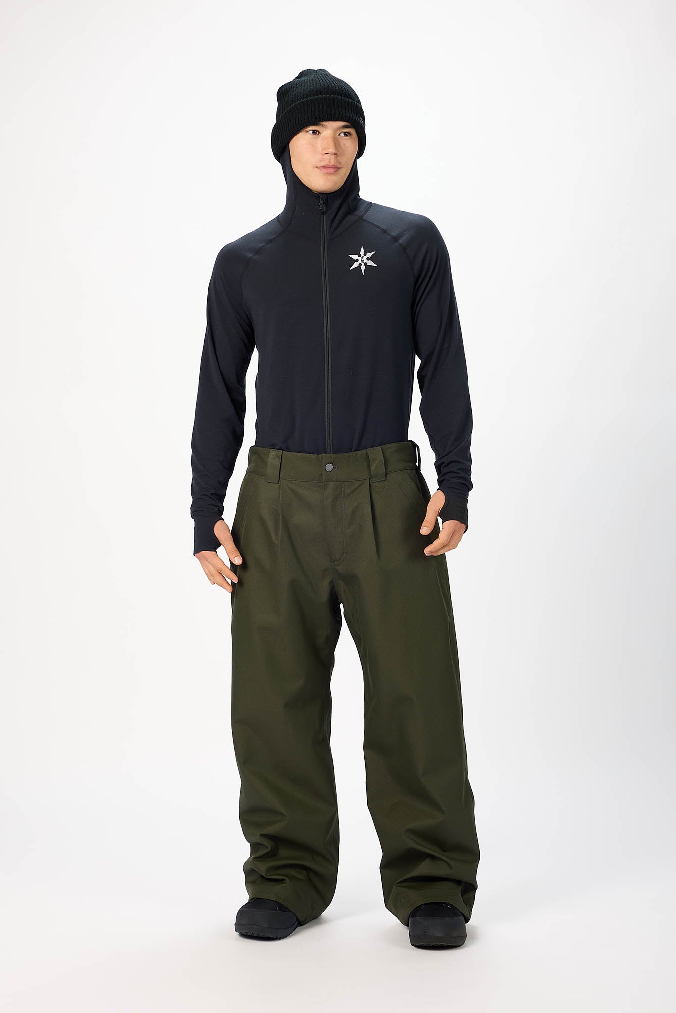 A model wearing a black long-sleeve athletic zip-up jacket with a white logo on the chest, olive green snow pants, a black beanie, and black boots, standing against a light gray studio background.