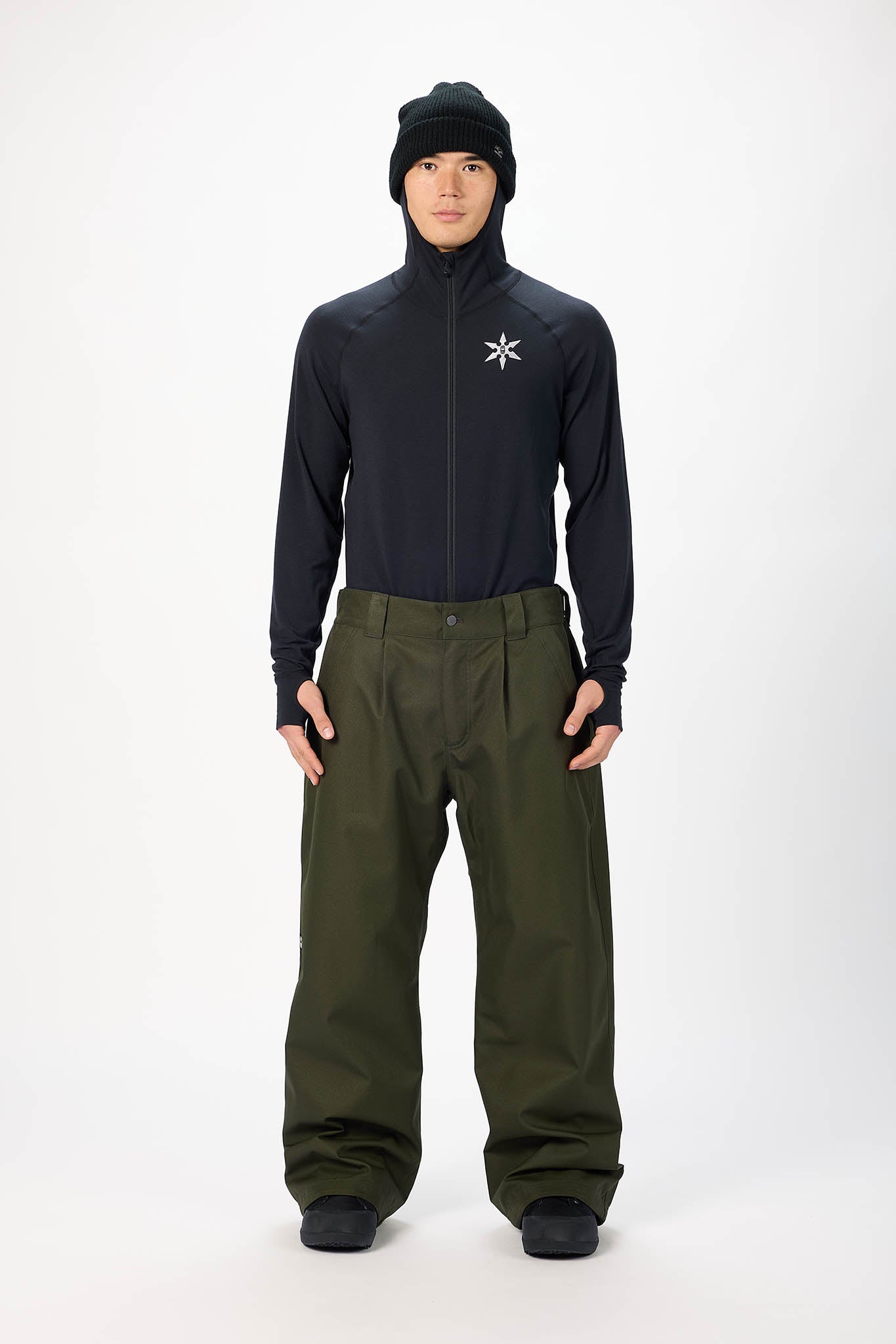 A male model wearing a black zip-up hooded jacket with a white star logo on the chest, paired with olive green snow pants, a black beanie, and black gloves, standing against a plain white studio background.