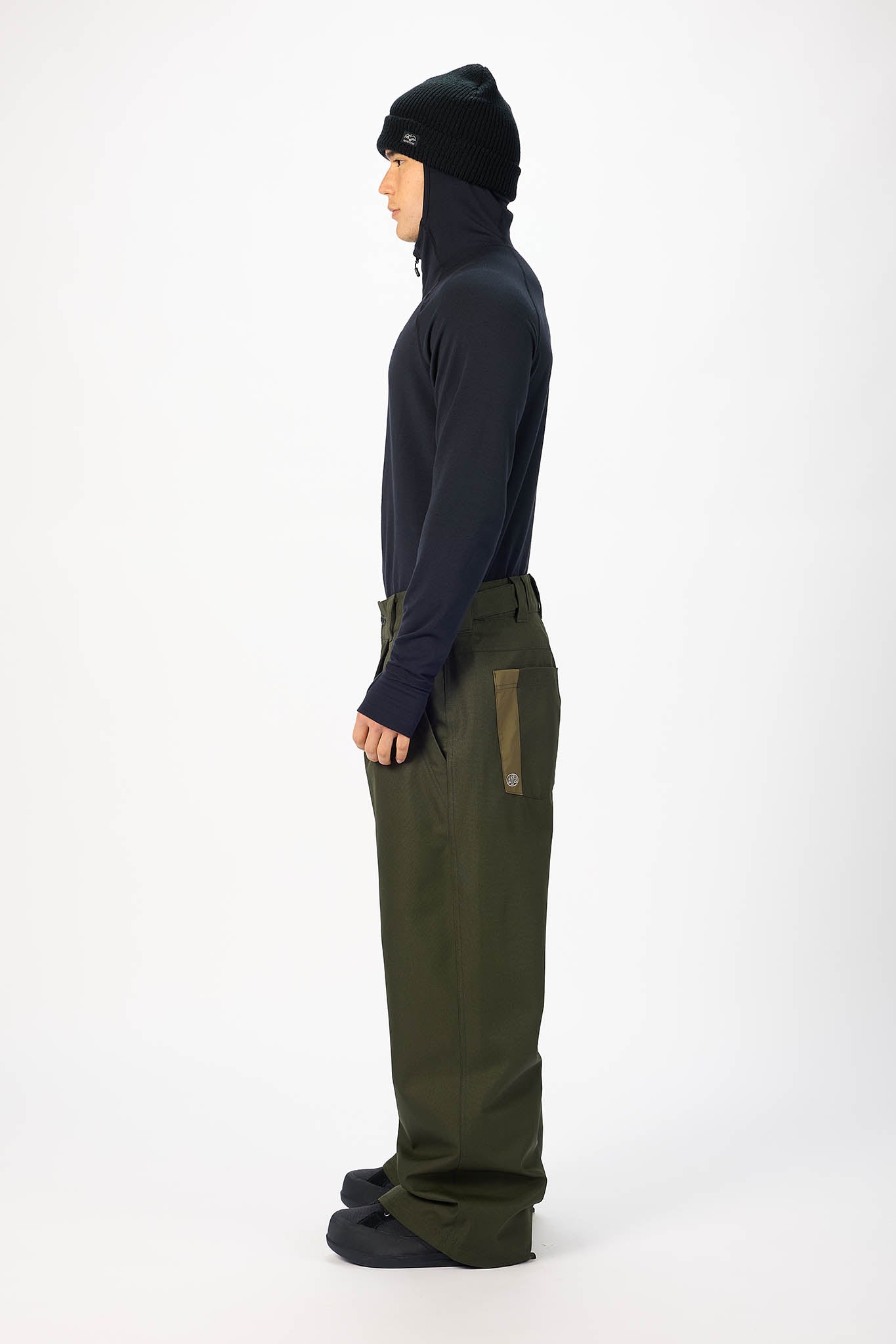 A male model wearing a black zip-up hooded jacket with a white star logo on the chest, paired with olive green snow pants, a black beanie, and black gloves, standing against a plain white studio background. In side profile