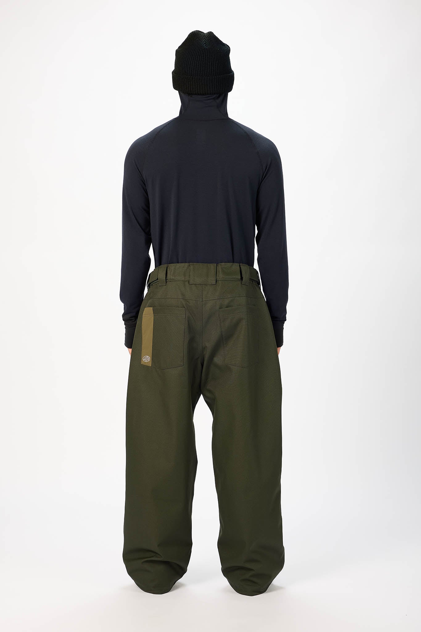 A model shown from the back wearing a black beanie, a dark navy long-sleeve top, and olive green wide-leg pants with a tan patch on the back pocket, set against a plain white studio background.