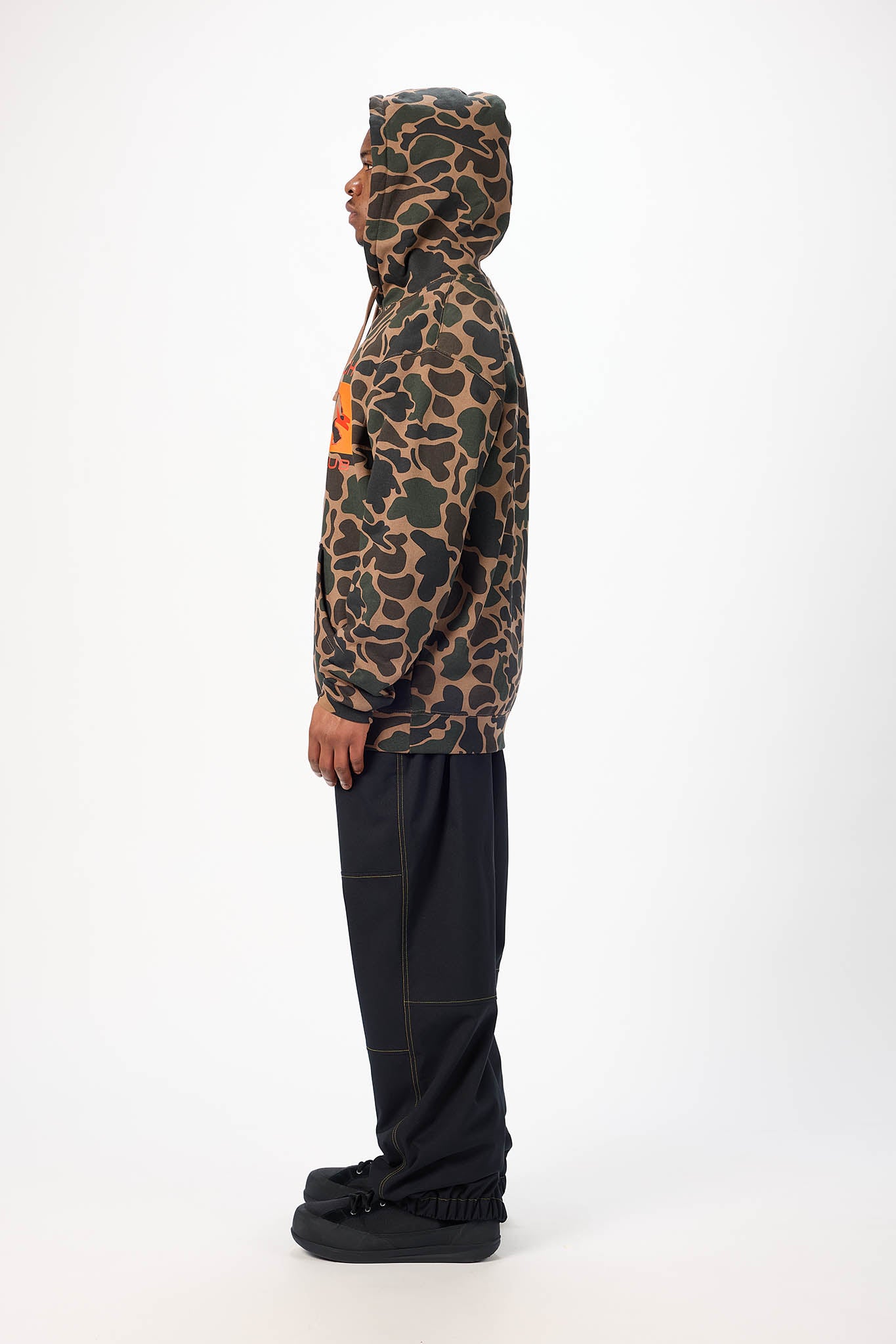 A man modeling a camouflage hoodie with a prominent orange graphic on the chest, paired with black pants and black sneakers, against a plain white studio background.