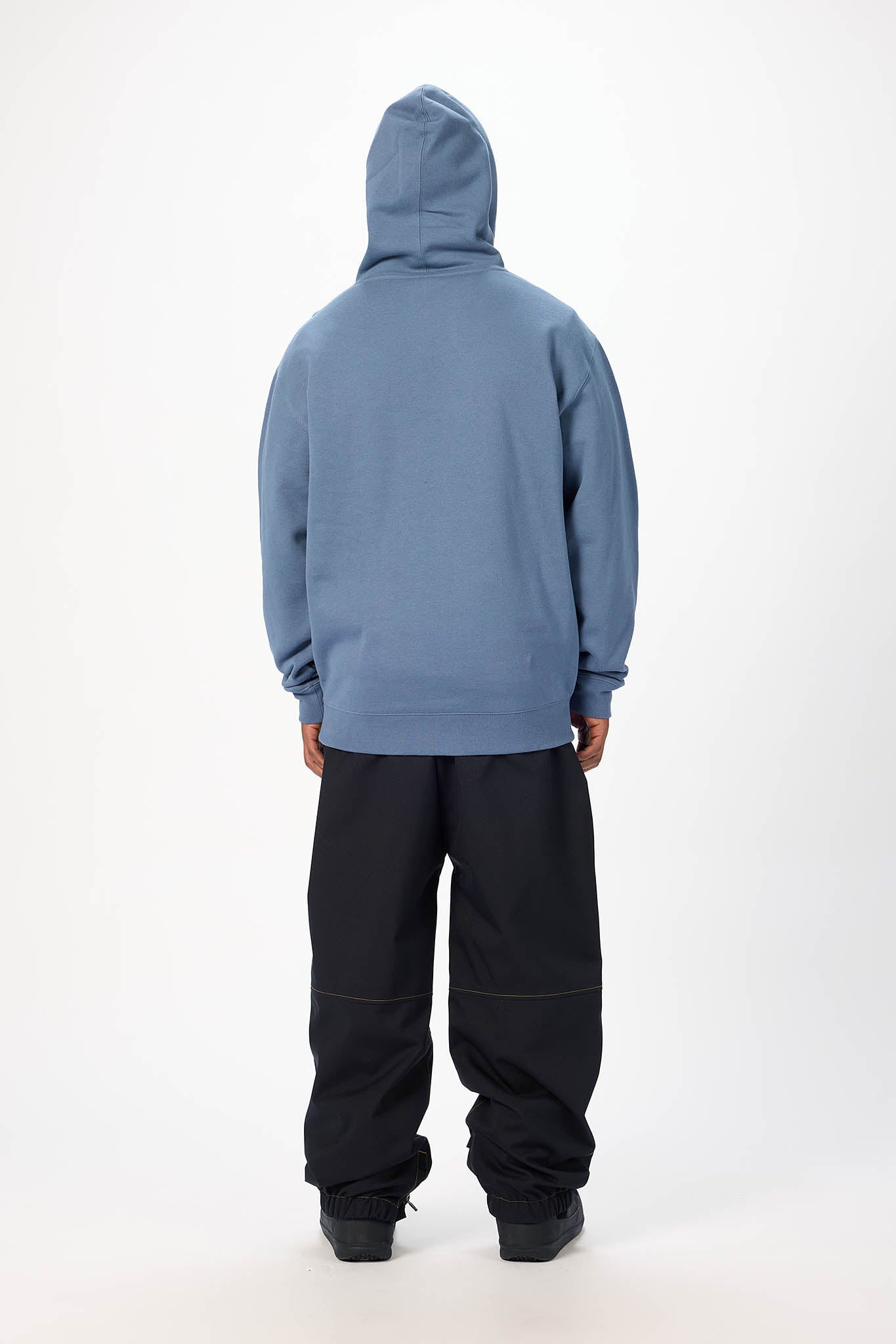 A man in profile wearing a dusty blue pullover hoodie with a white chest patch, black pants, and black sneakers, standing against a plain white studio background. viewed from behind