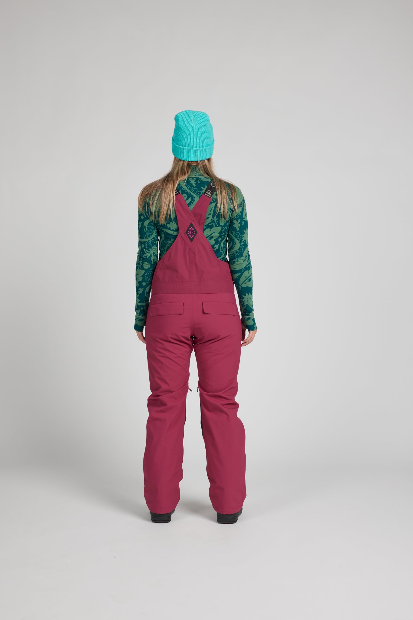 A model viewed from the back wearing a bright turquoise beanie, a green and teal patterned long-sleeve shirt, and magenta snow pants over a light gray studio background.