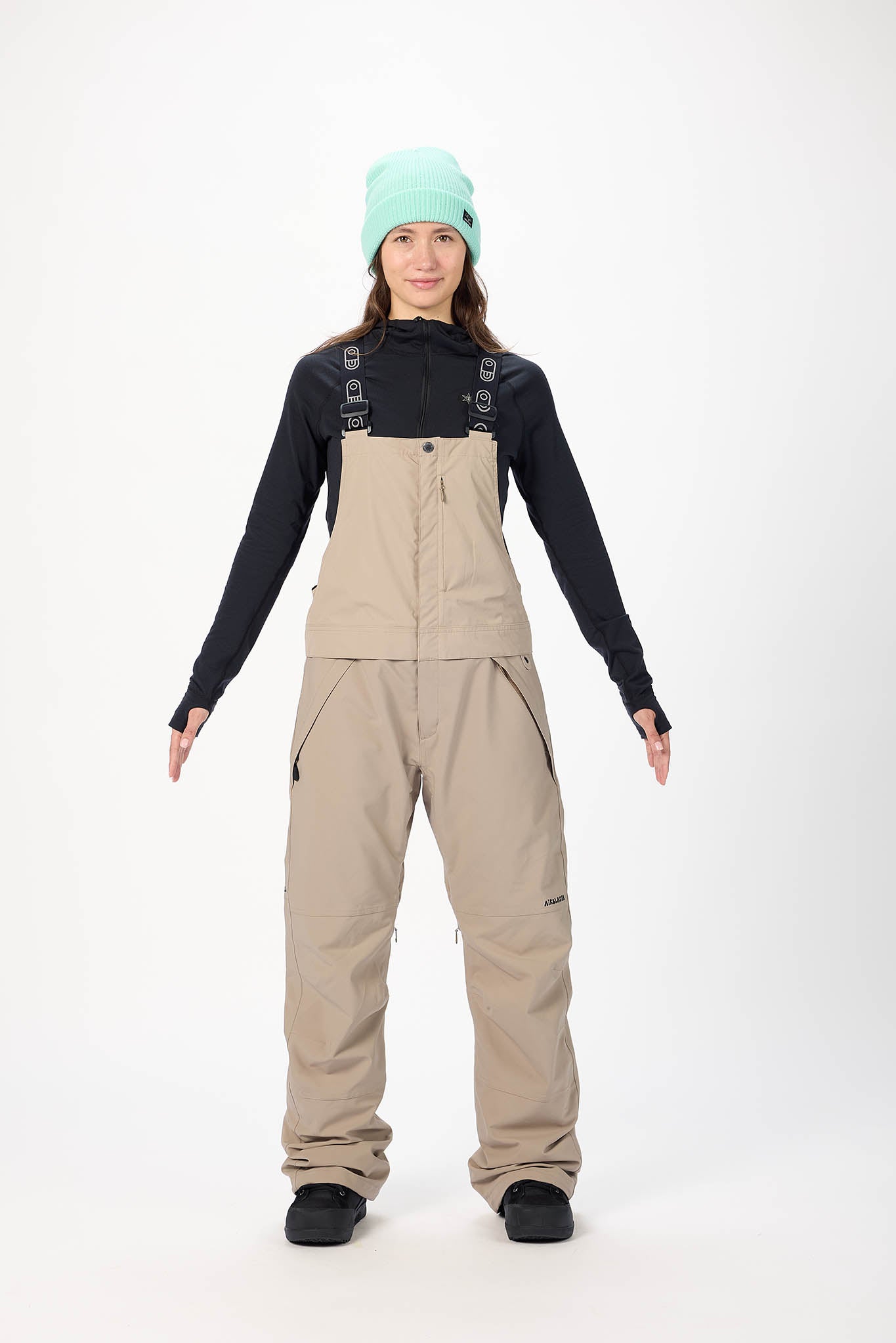 Person wearing a black long-sleeve shirt, beige bib overalls, and a light green beanie on a white background