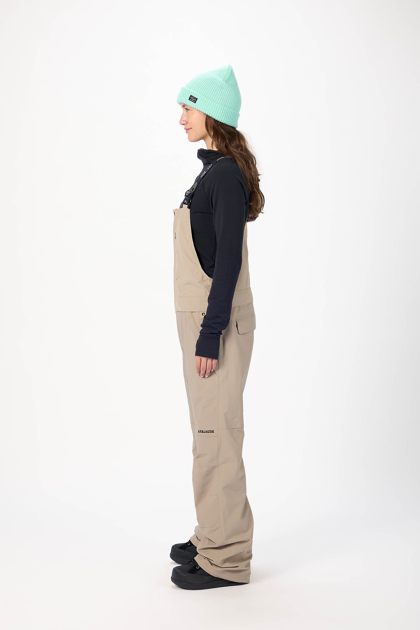 Person wearing a dark jacket, beige bib overalls, and a light green beanie on a white background