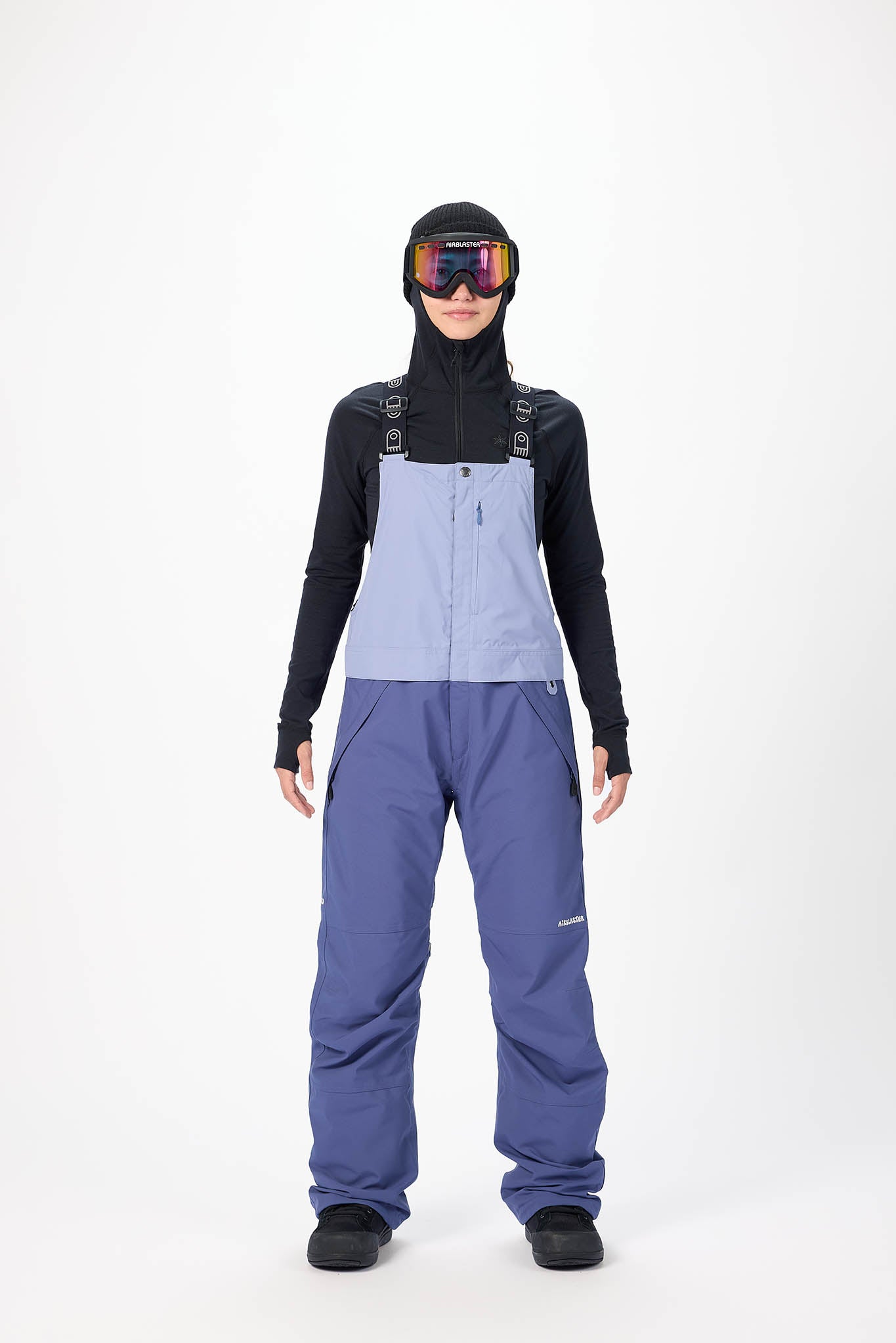 Person wearing a black jacket, blue bib overalls, and goggles on a white background