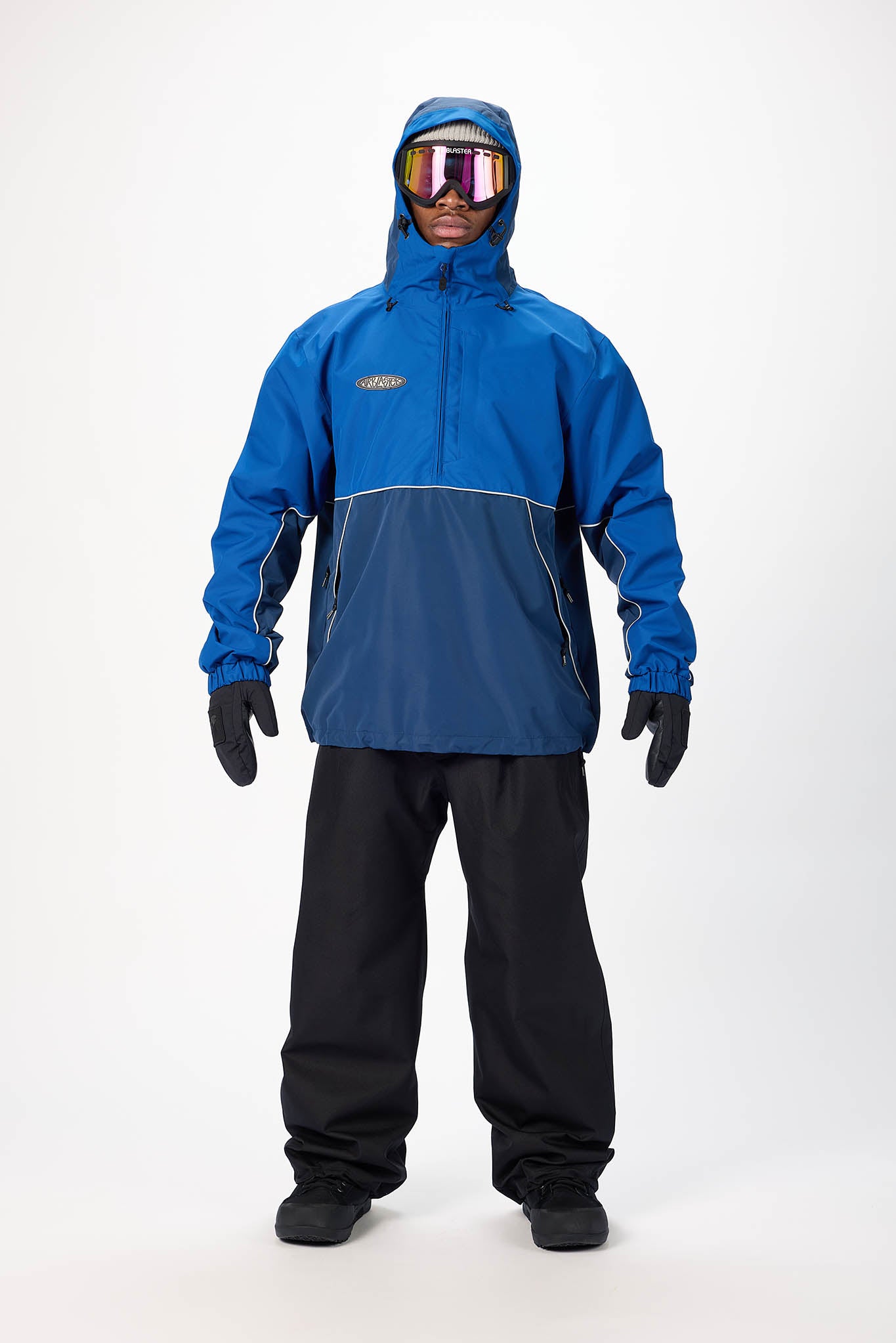 A model wearing a blue and navy colorblock snow jacket with a matching hood, black snow pants, black gloves, and reflective ski goggles, standing against a white studio background.