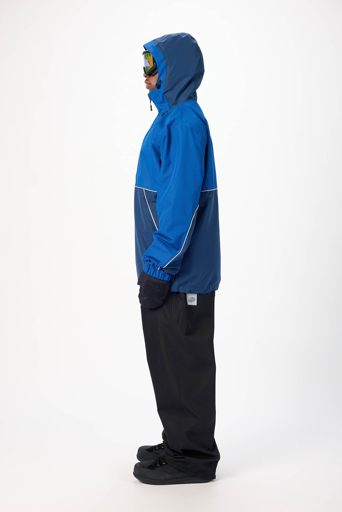 A person shown in side profile wearing a blue hooded jacket with dark pants and black snow boots, standing against a plain white studio background.