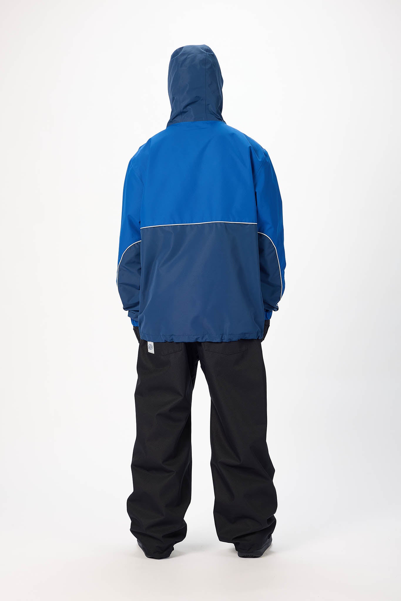A person shown from the back wearing a colorblock hooded jacket with a blue hood and upper back, a darker blue lower back, and black pants, photographed against a plain white studio background.