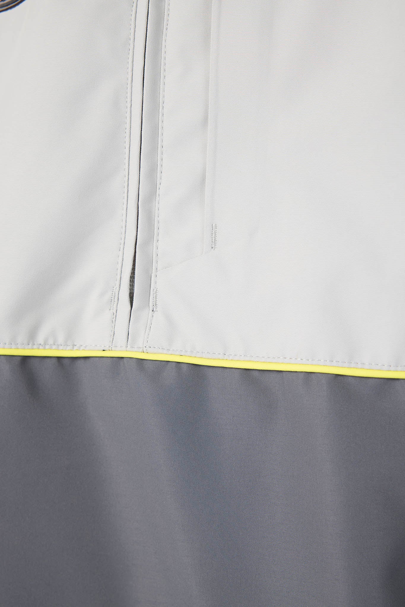 Close-up detail of a lightweight jacket showing a light gray upper section with a vertical zip pocket, a darker gray lower section, and a thin bright yellow piping accent.