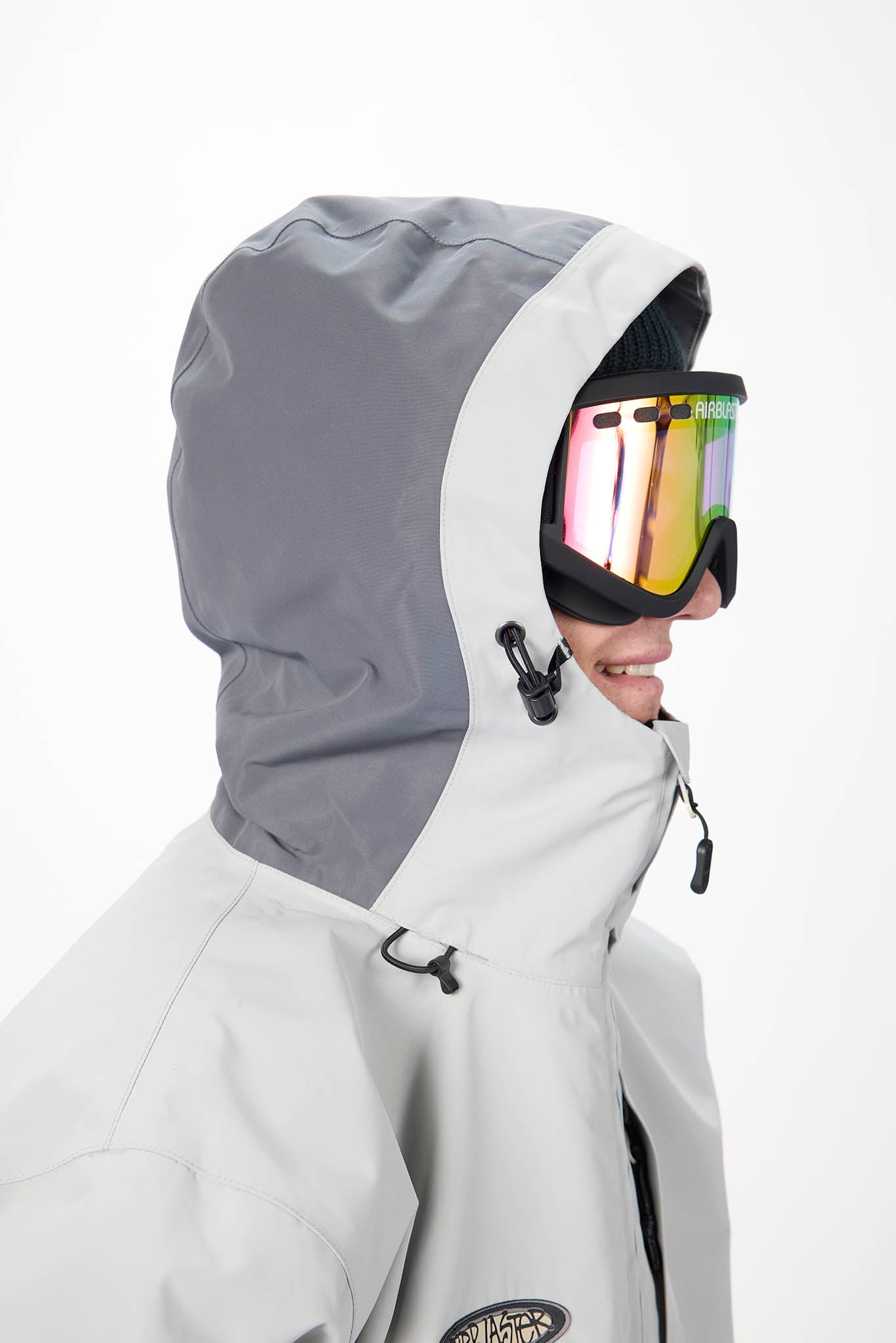 A close-up side profile of a person wearing a light gray and dark gray ski jacket with a matching hood, black goggles with a reflective gold and pink lens, and a black beanie against a plain white background.