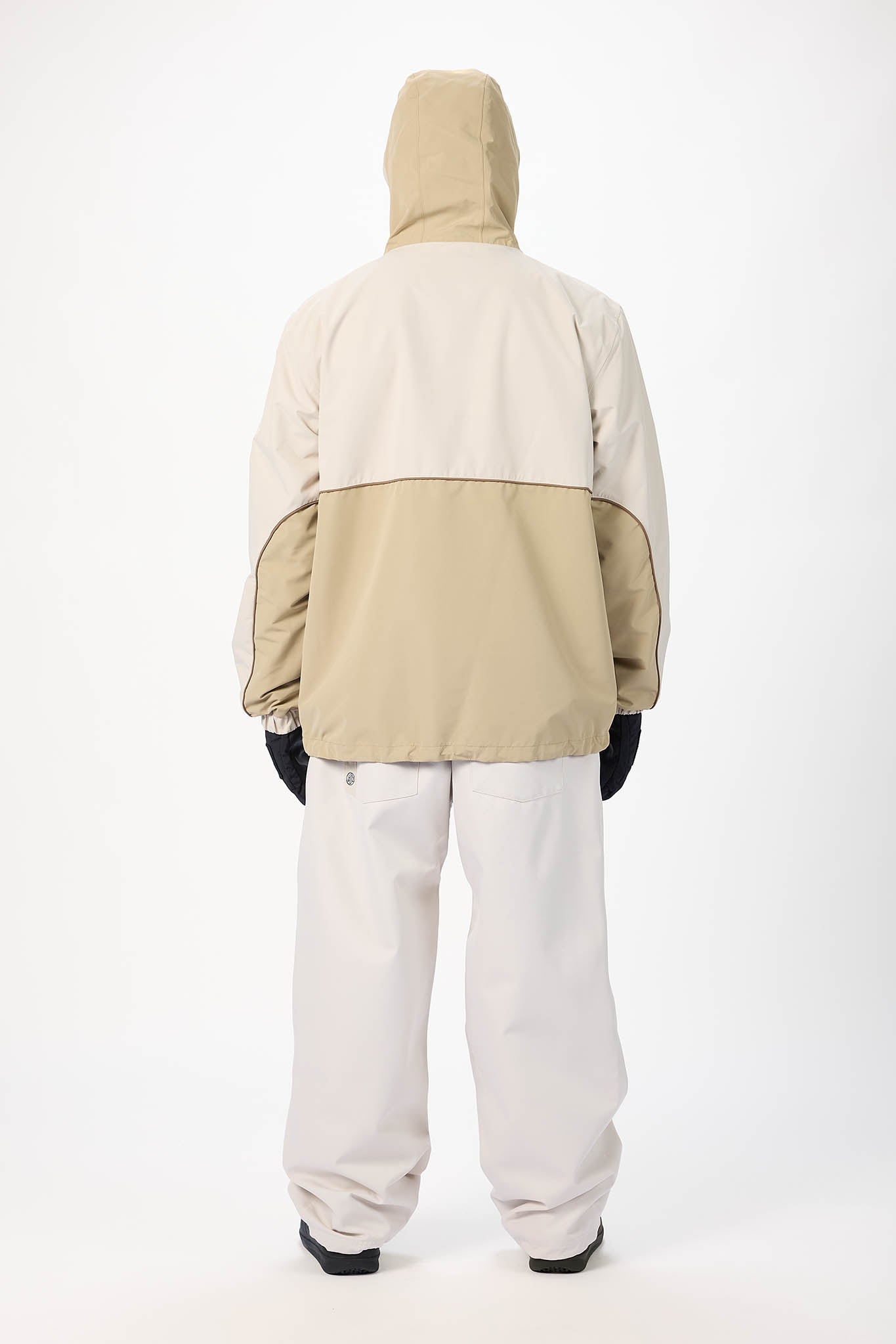 A person shown from the back wearing a color-blocked hooded jacket with a beige hood and lower panel, a light beige upper section, and light gray pants, standing against a plain white studio background.