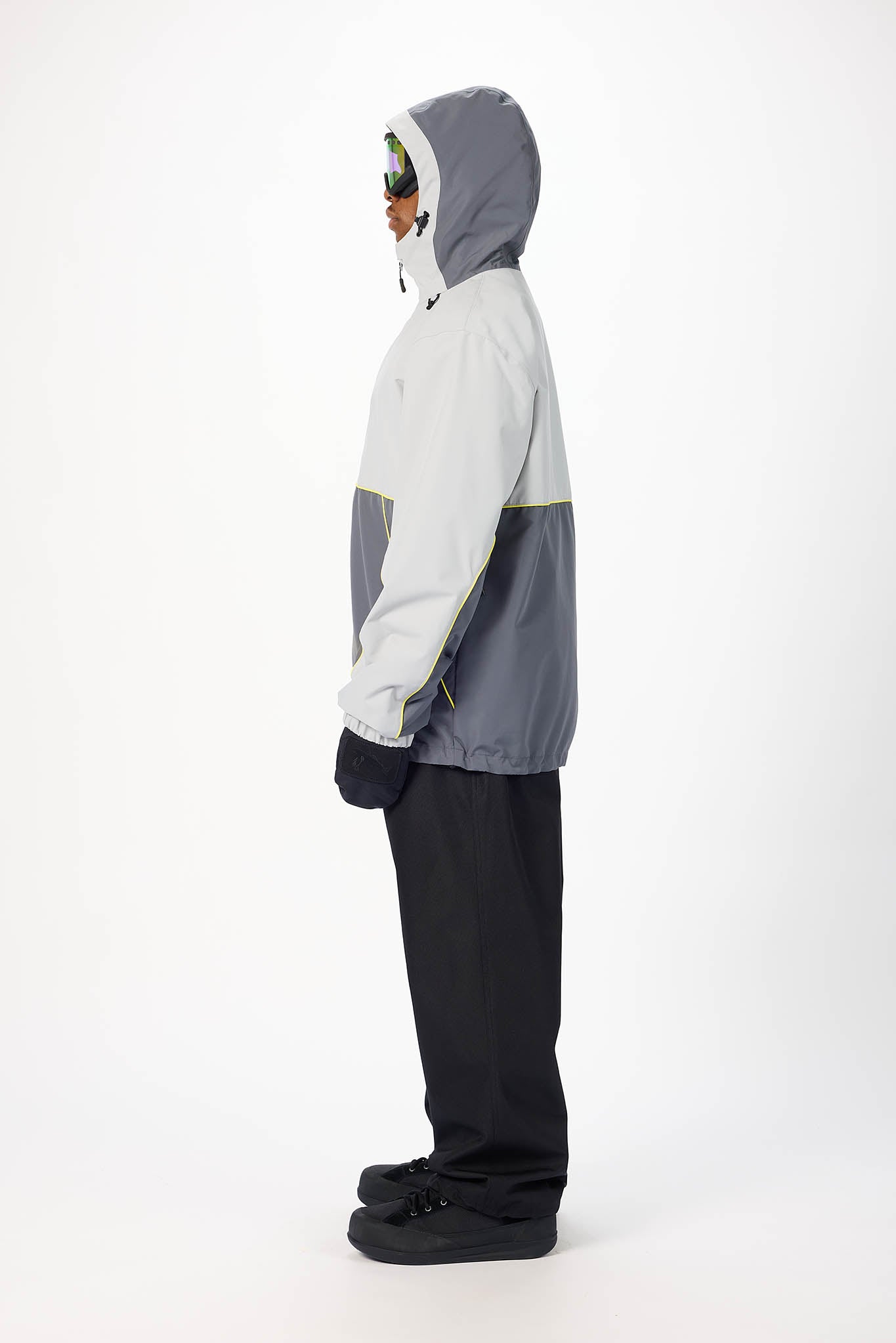 A person shown in profile wearing a color-blocked hooded jacket with a light gray upper section, dark gray lower section, and black pants against a plain white studio background.