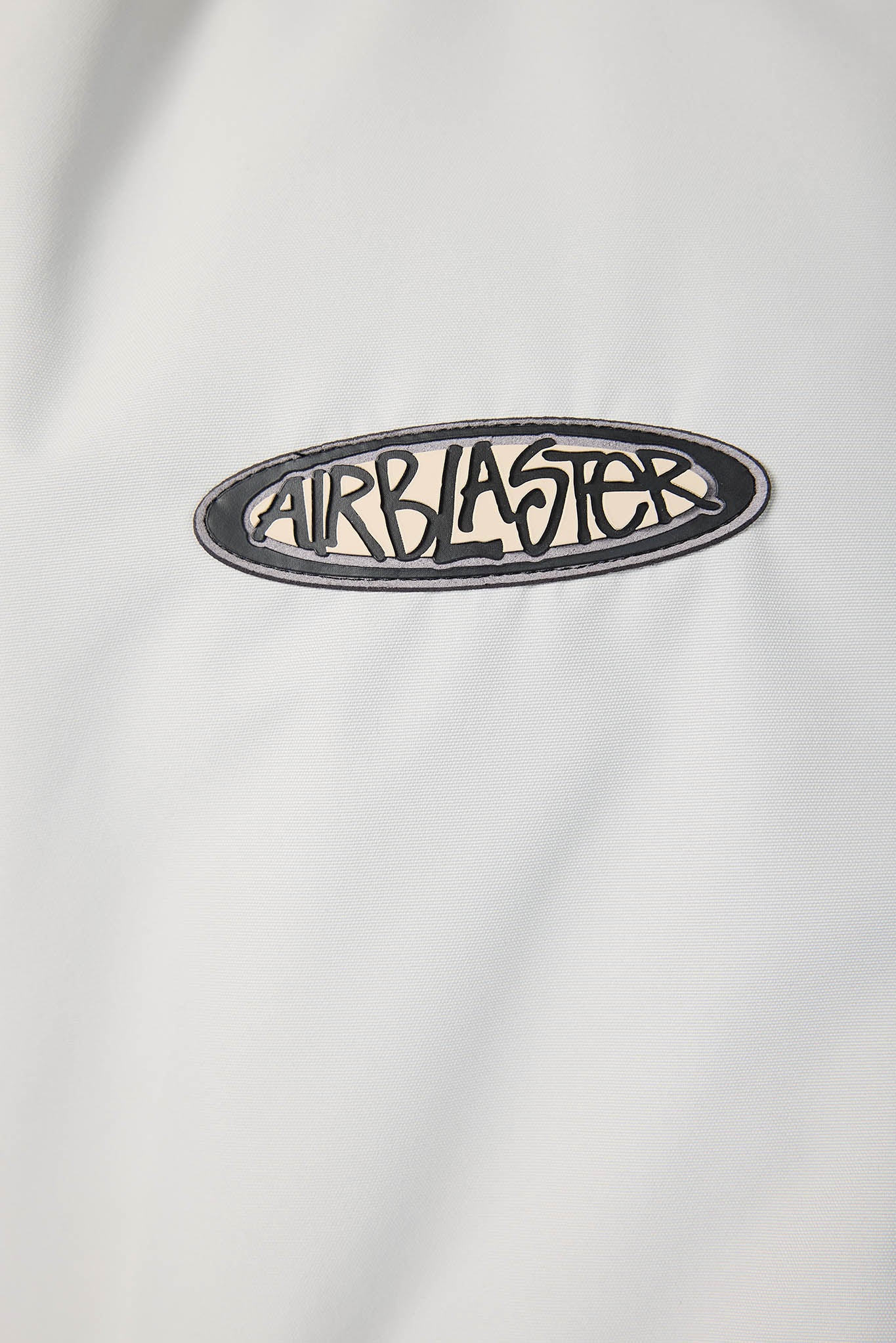 A close-up of a white garment featuring an oval embroidered patch with the brand name 'AIRBLASTER' in black stylized lettering.