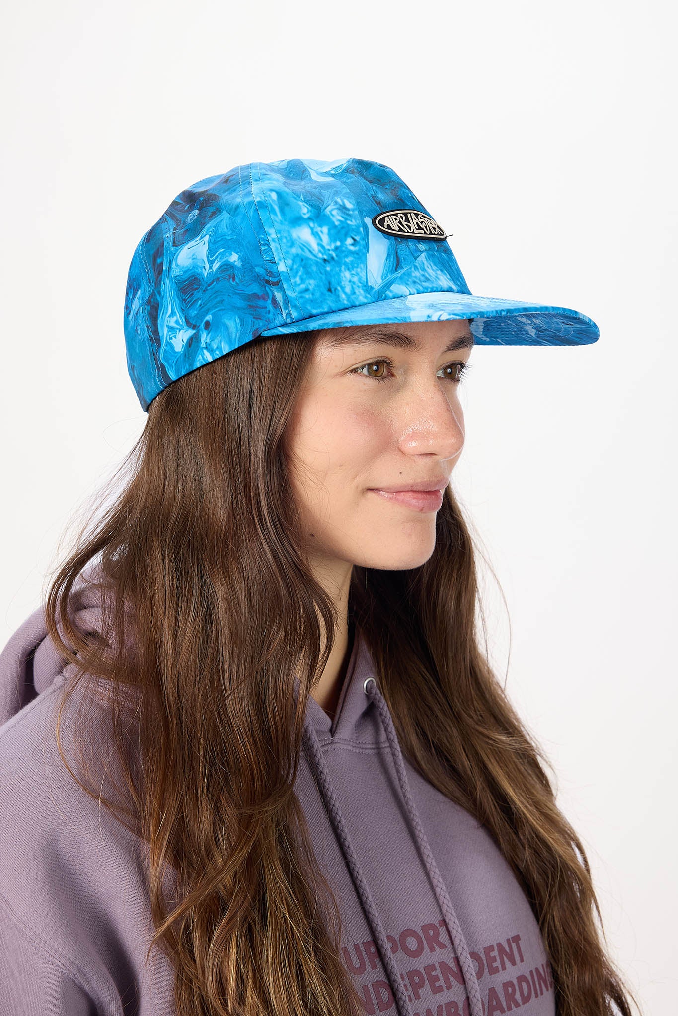 A woman with long brown hair wearing a blue marbled patterned baseball cap and a lavender hoodie, shown in profile against a plain white background.