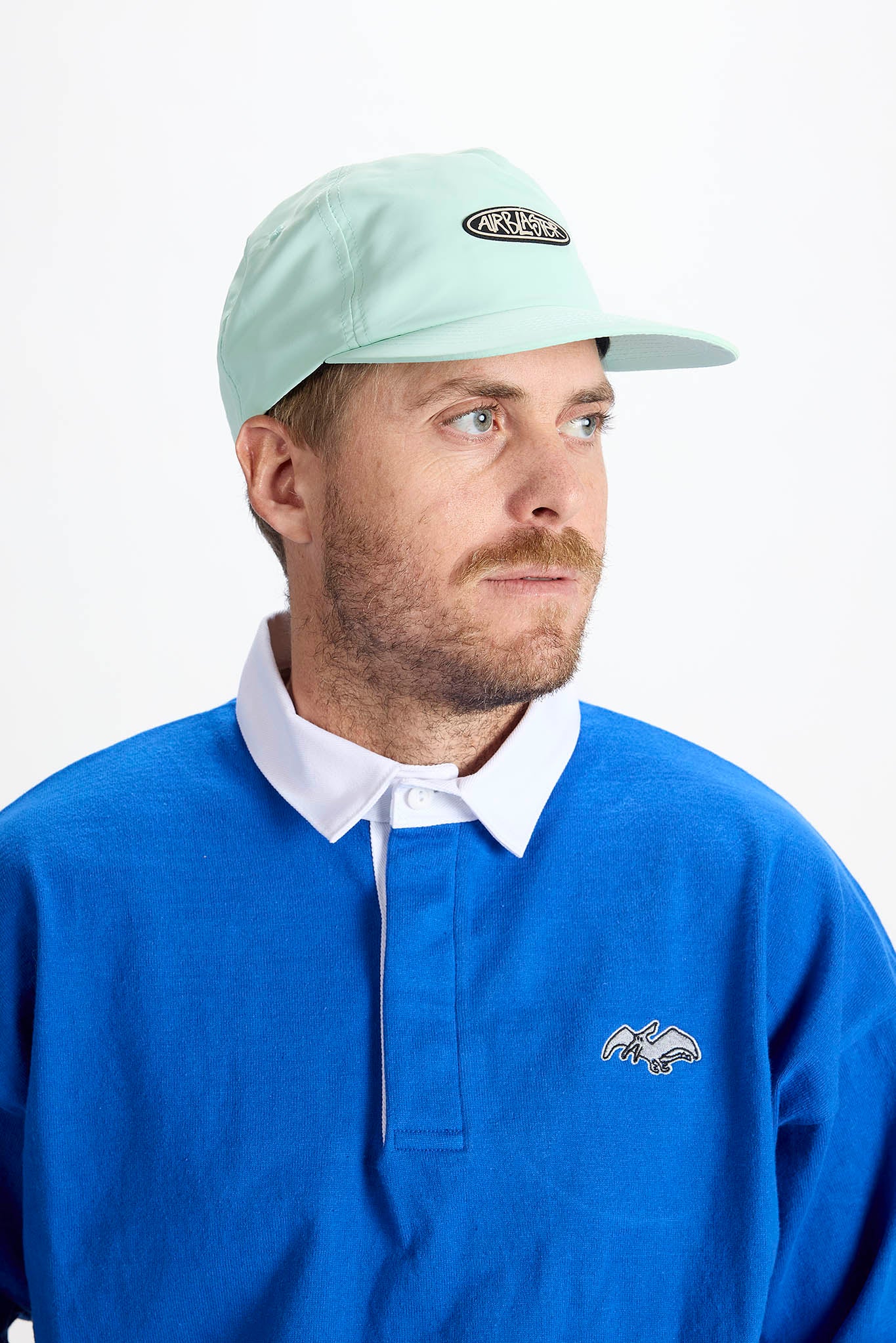 A man wearing a light mint green baseball cap with a black oval logo and a royal blue polo shirt with a white collar, photographed against a plain white background.