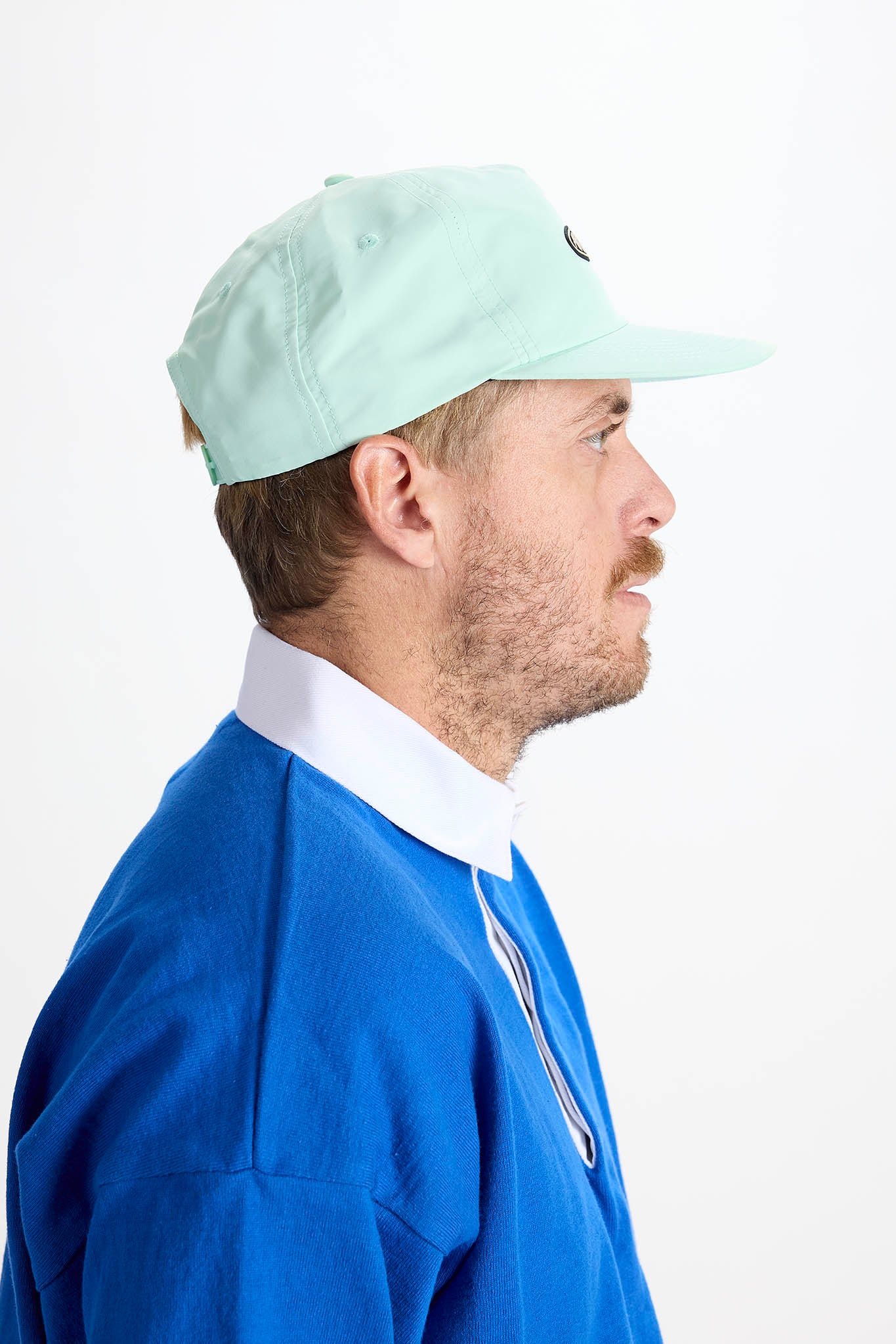 side profile A man wearing a light mint green baseball cap with a black oval logo and a royal blue polo shirt with a white collar, photographed against a plain white background.