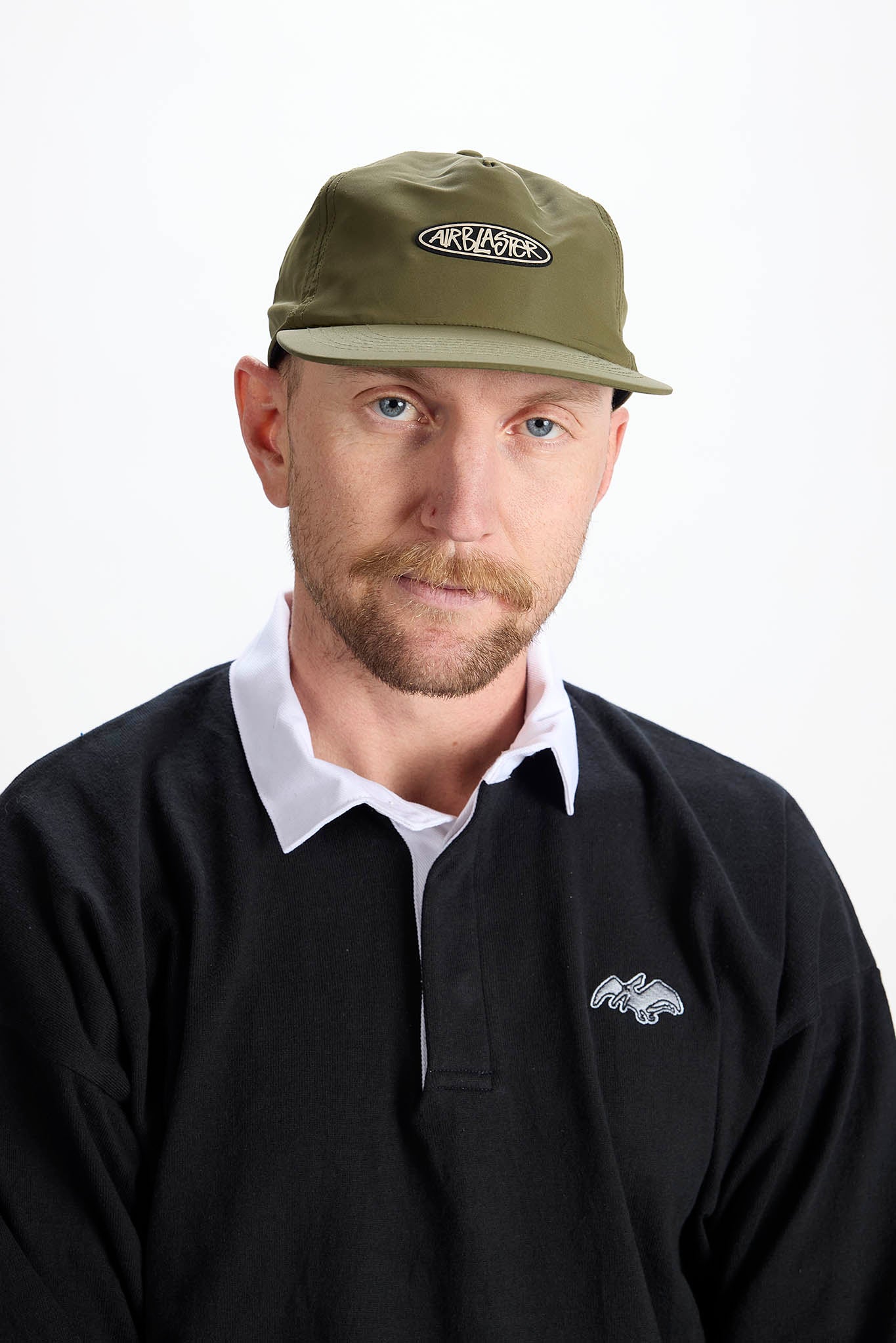 A man wearing an olive green baseball cap with a white embroidered logo and a black long-sleeve polo shirt over a white collared shirt, photographed against a plain white background.