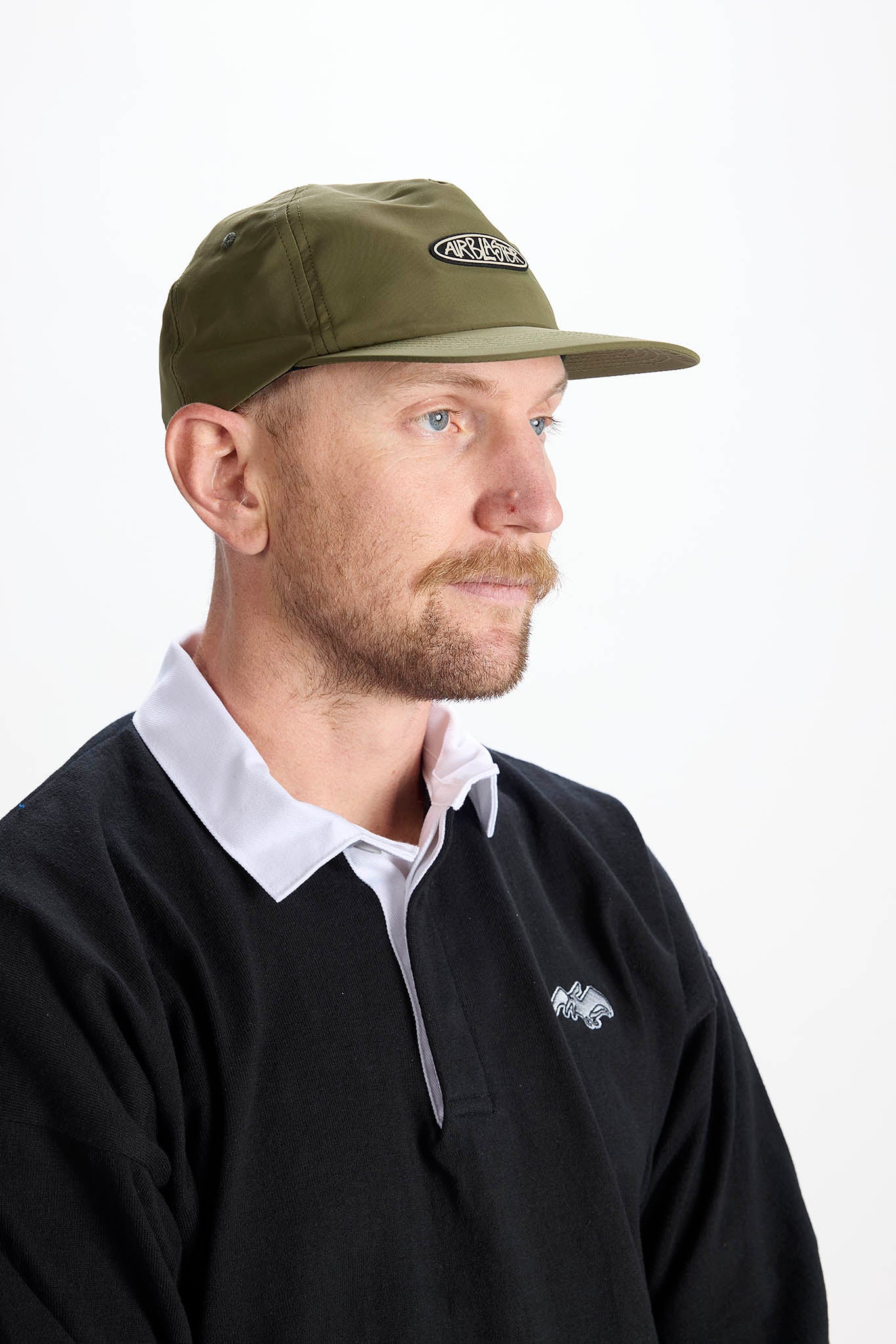 A man wearing an olive green baseball cap with a white embroidered logo and a black long-sleeve polo shirt over a white collared shirt, photographed against a plain white background.