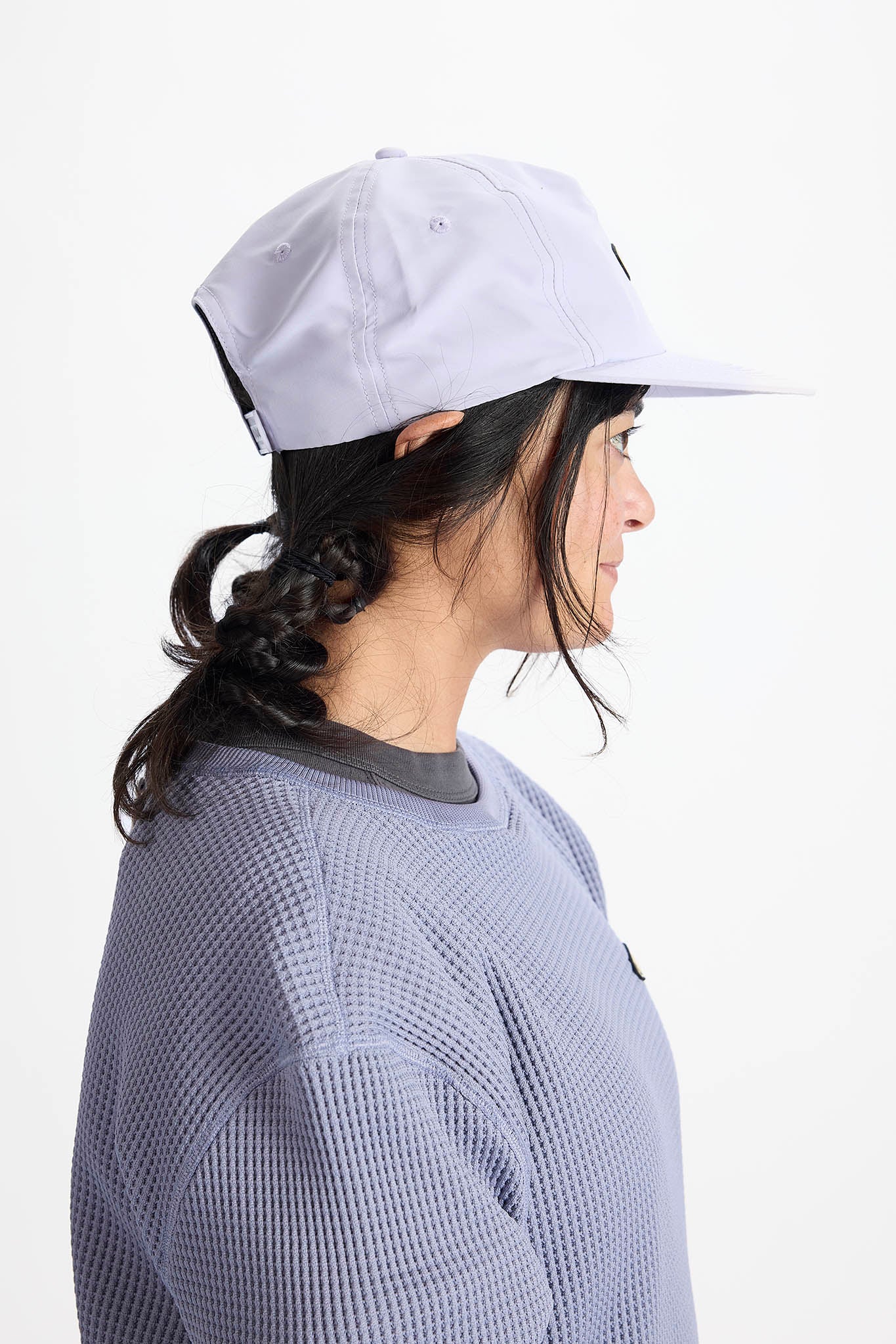 A side-profile view of a woman wearing a light gray baseball cap and a textured lavender-gray long-sleeve shirt against a plain white background.