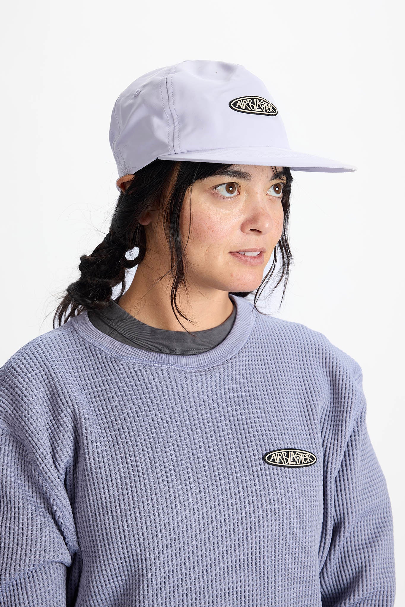 A woman wearing a light lavender waffle-knit cap and matching waffle-knit sweatshirt, both featuring an oval black and white logo on the front, against a plain white background.